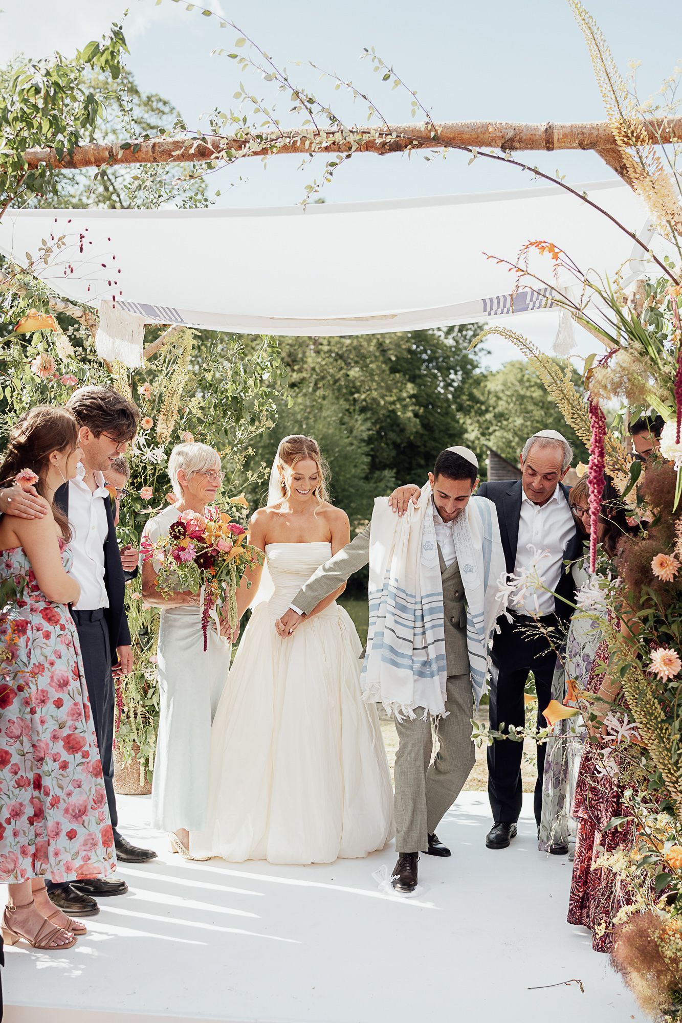 Jewish wedding ceremony outdoors under a floral chuppah, bride in white gown with bouquet, groom in tallit breaking glass.