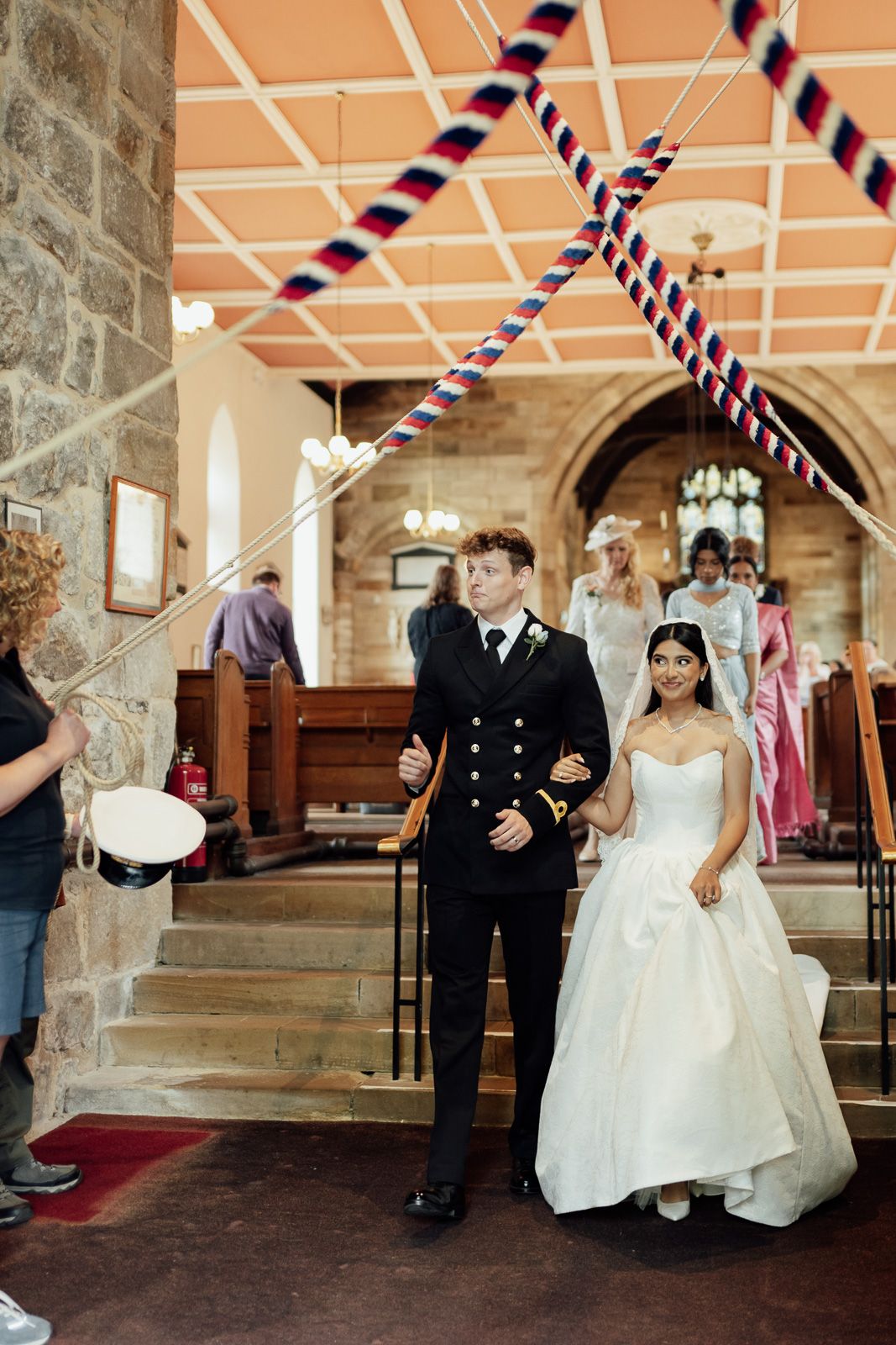 Bride and groom walking down church aisle, groom in naval uniform, bride in white gown, colorful ribbon arch overhead.