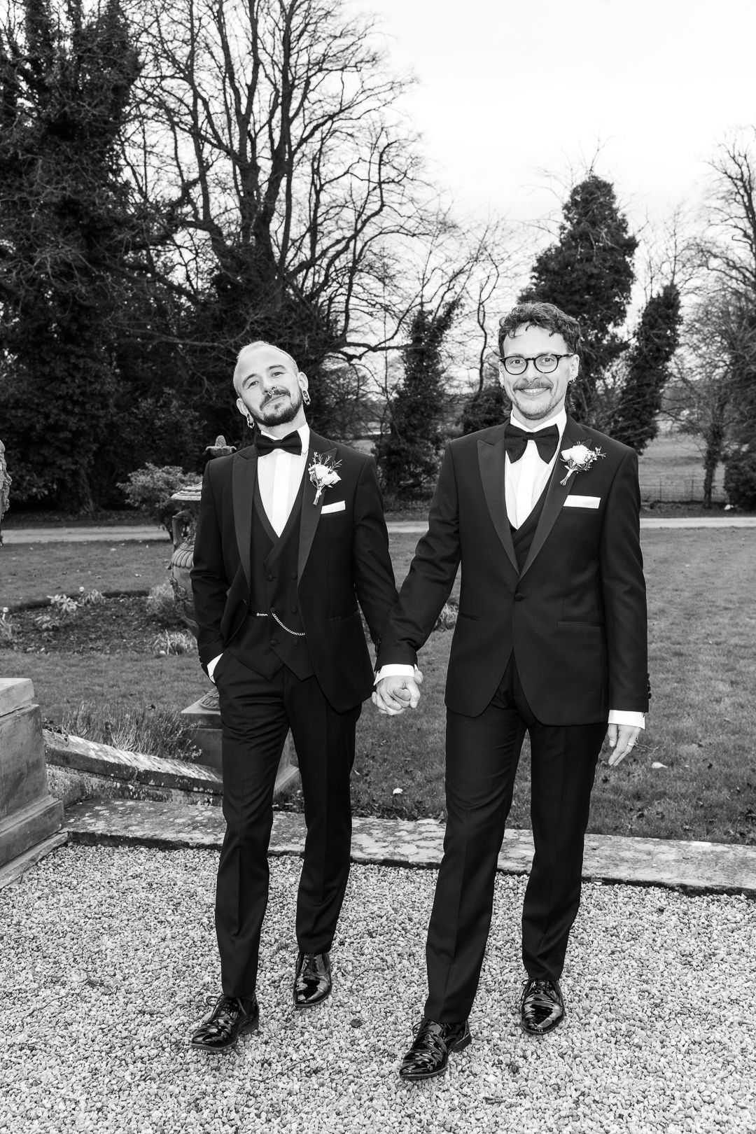 Two people in black tuxedos with bow ties hold hands while standing on gravel path in garden setting.