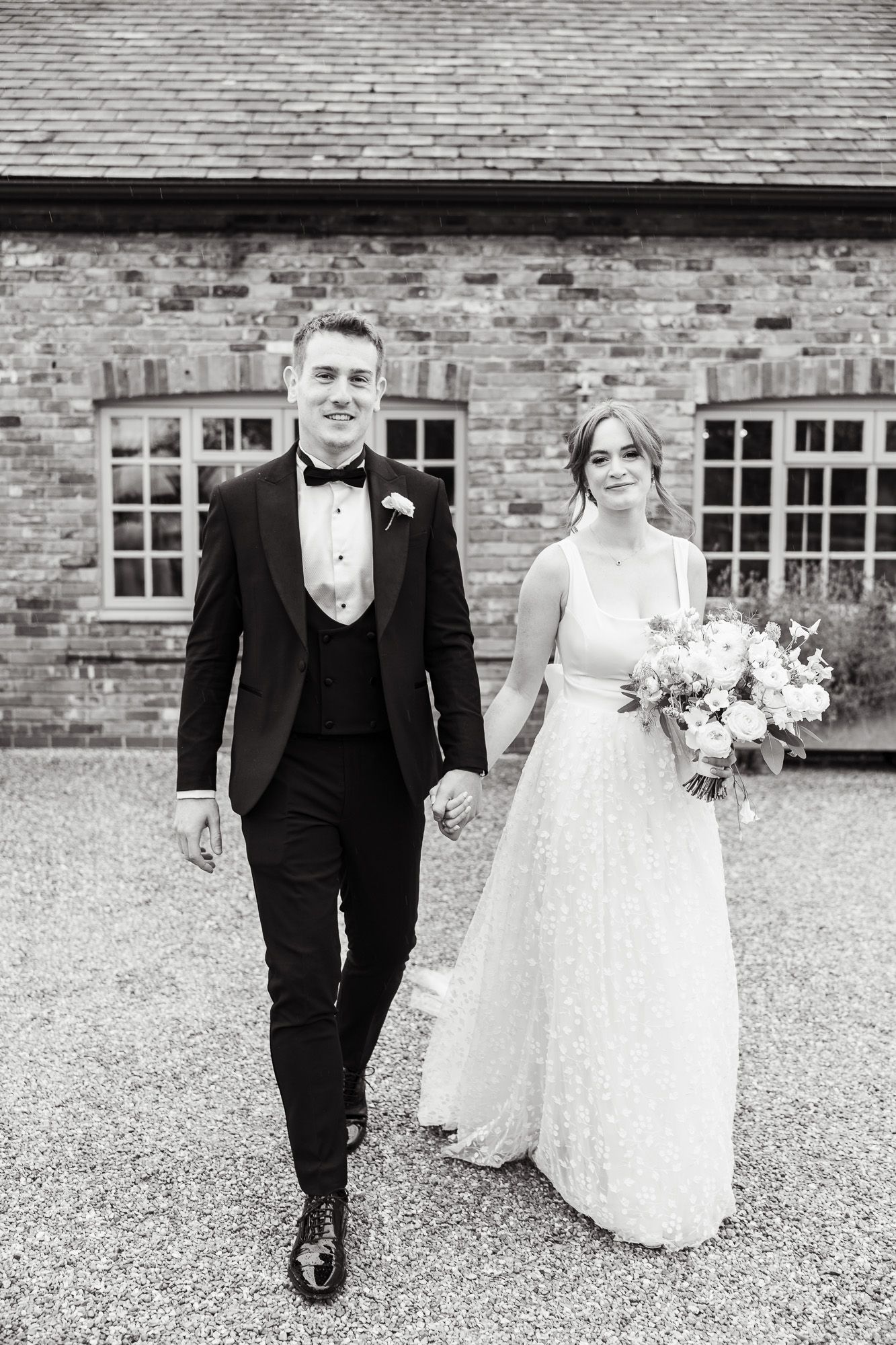 Black and white wedding photo showing couple walking together outside brick building with bride holding floral bouquet.