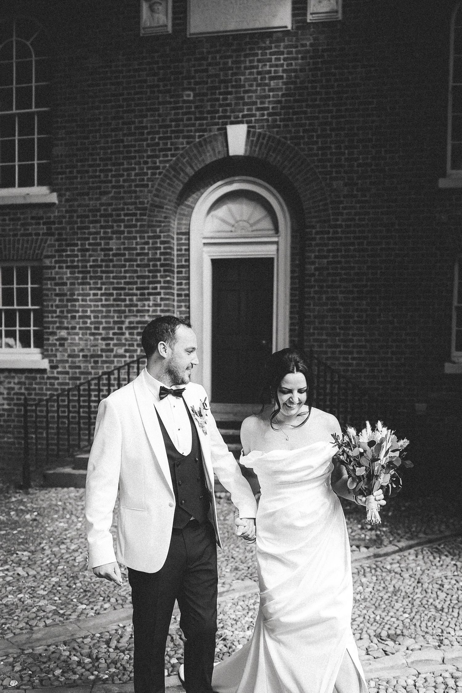 A bride and her companion share a candid moment while walking on a cobblestone path outside a brick building.