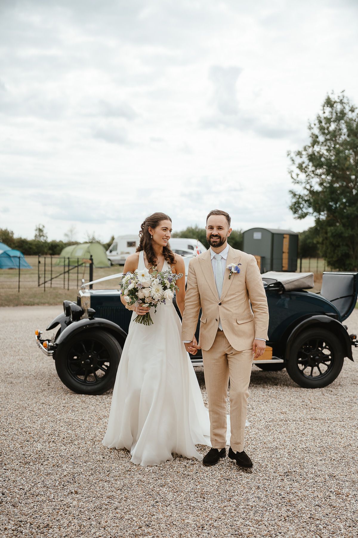 Happy couple laughs together in front of vintage car on their wedding day wearing beige suit and white gown.