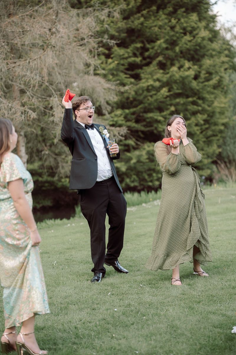 Joyful wedding guests in formal attire dance and celebrate outdoors on a grassy lawn with evergreen trees in background.