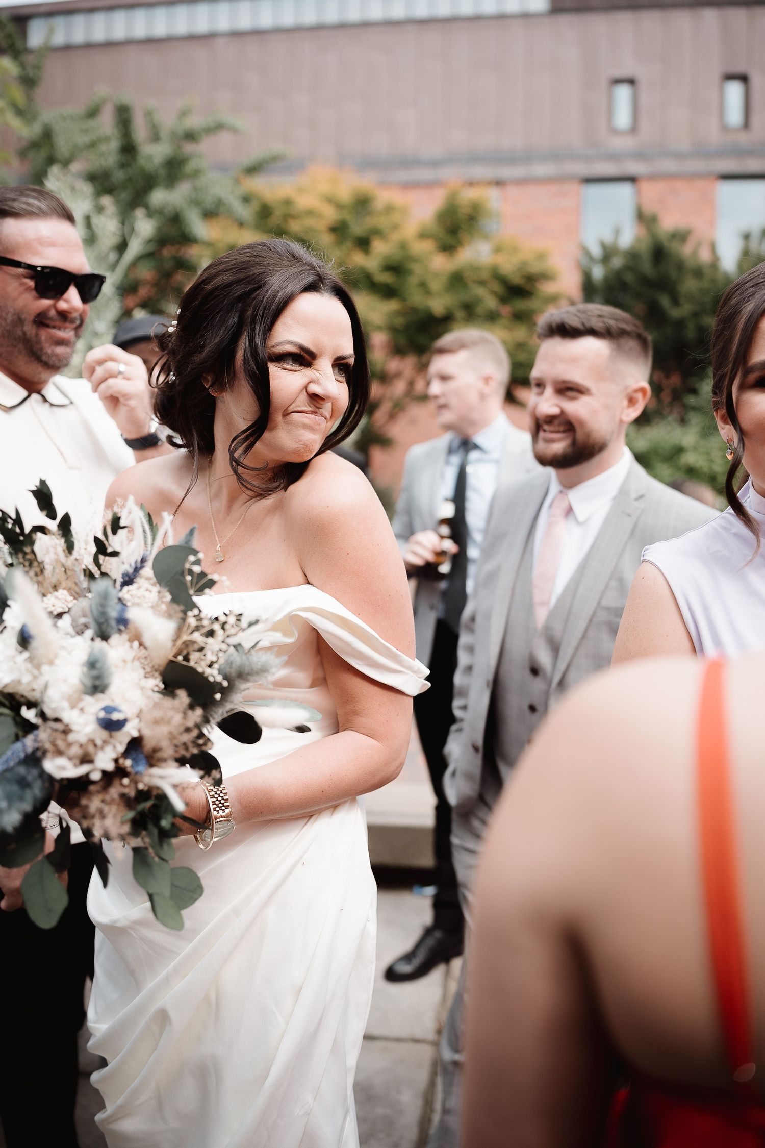 Wedding guests gather outdoors while bride in off-shoulder white gown holds beautiful eucalyptus and floral bouquet.