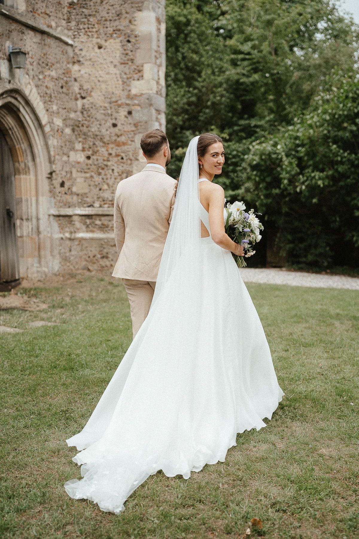 Bride in flowing white wedding dress holding bouquet stands with groom near stone church wall in garden setting.