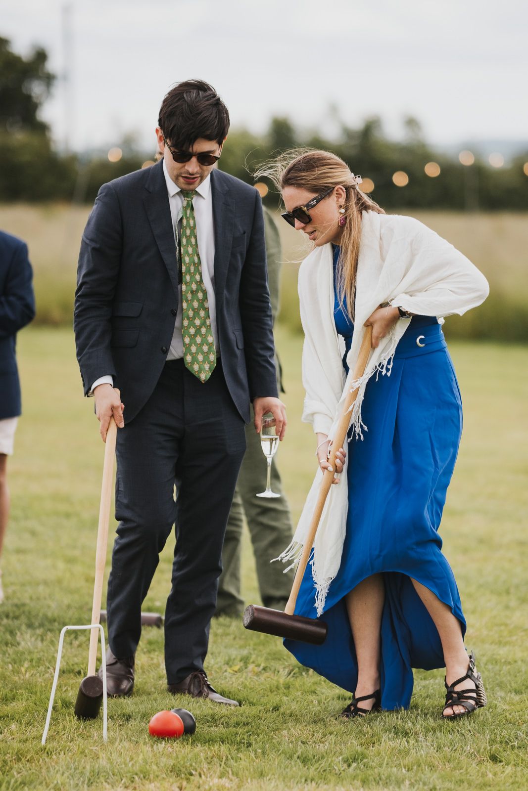 Two people dressed in formal attire play croquet on a lawn during what appears to be an outdoor event.