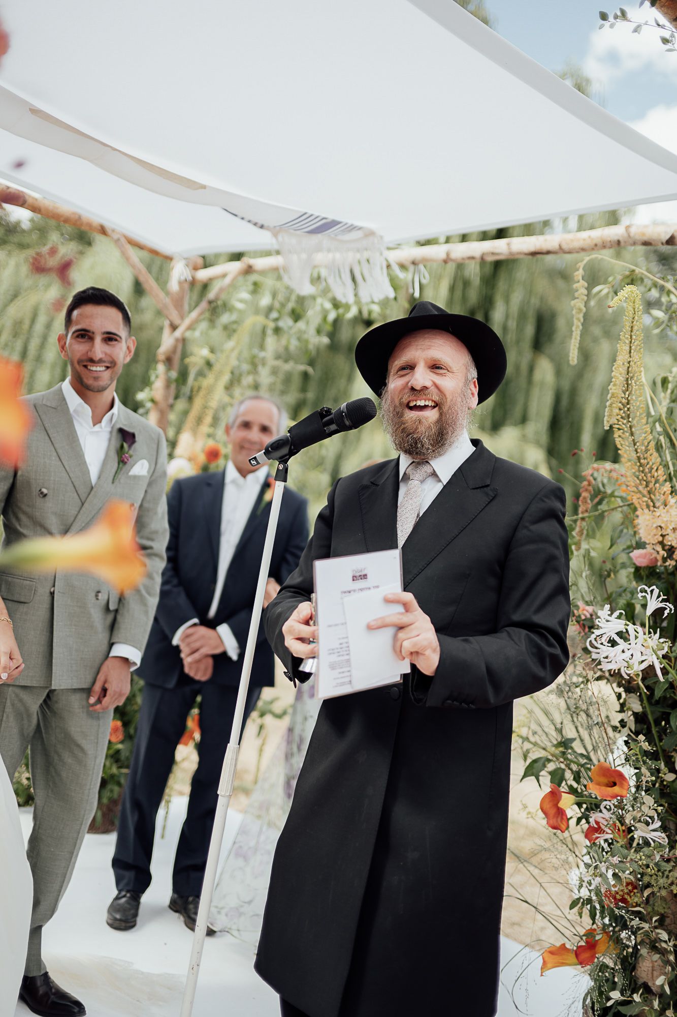 Rabbi officiates Jewish wedding ceremony under chuppah, holding ketubah, with smiling groom in gray suit standing nearby.