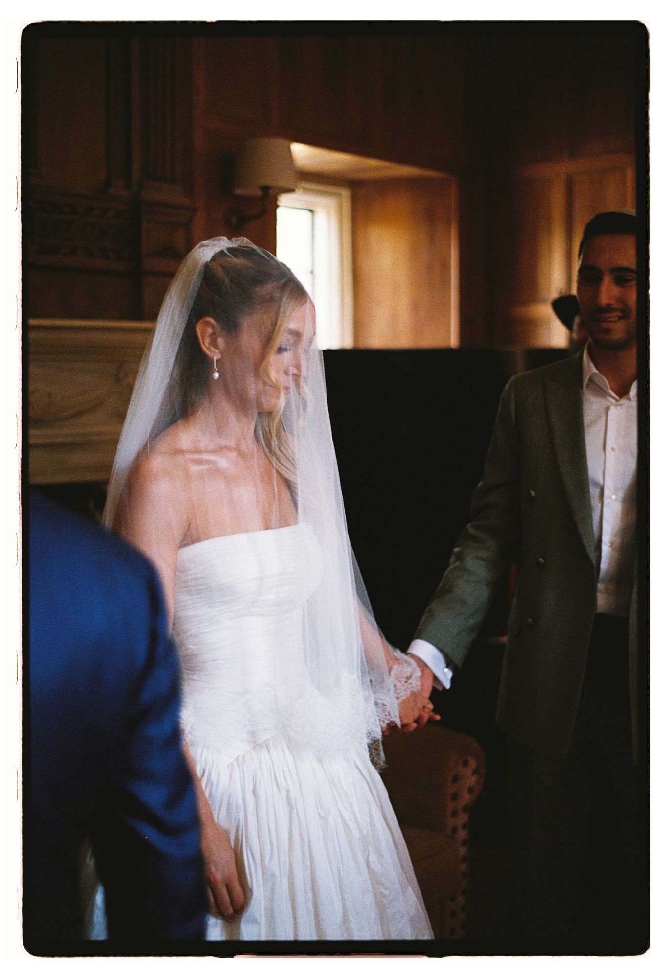 Bride in white strapless gown and veil holds hands with groom in dark suit during elegant indoor wedding ceremony.