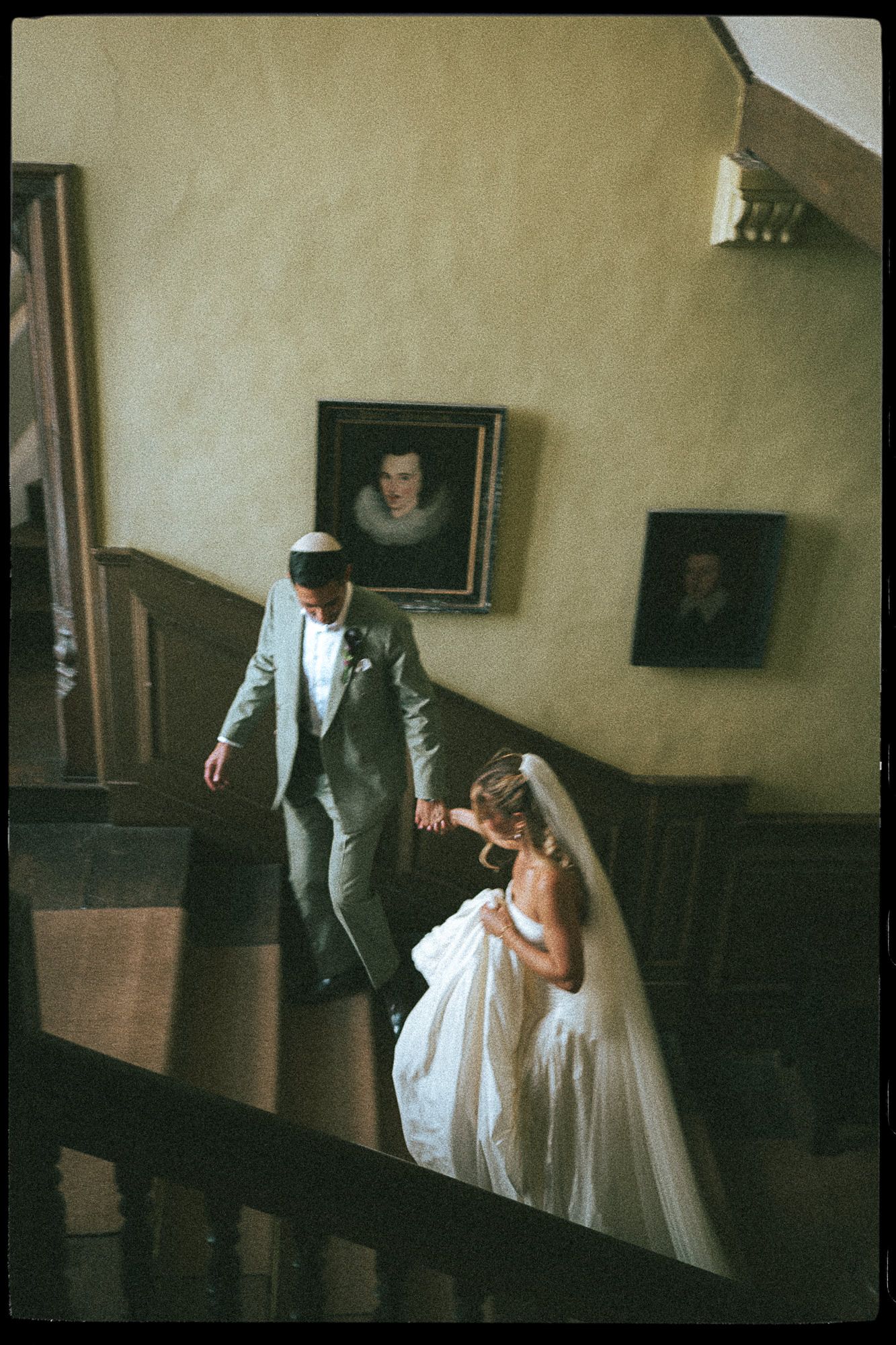 Jewish bride and groom descend grand staircase in elegant venue, with portrait paintings on wall.