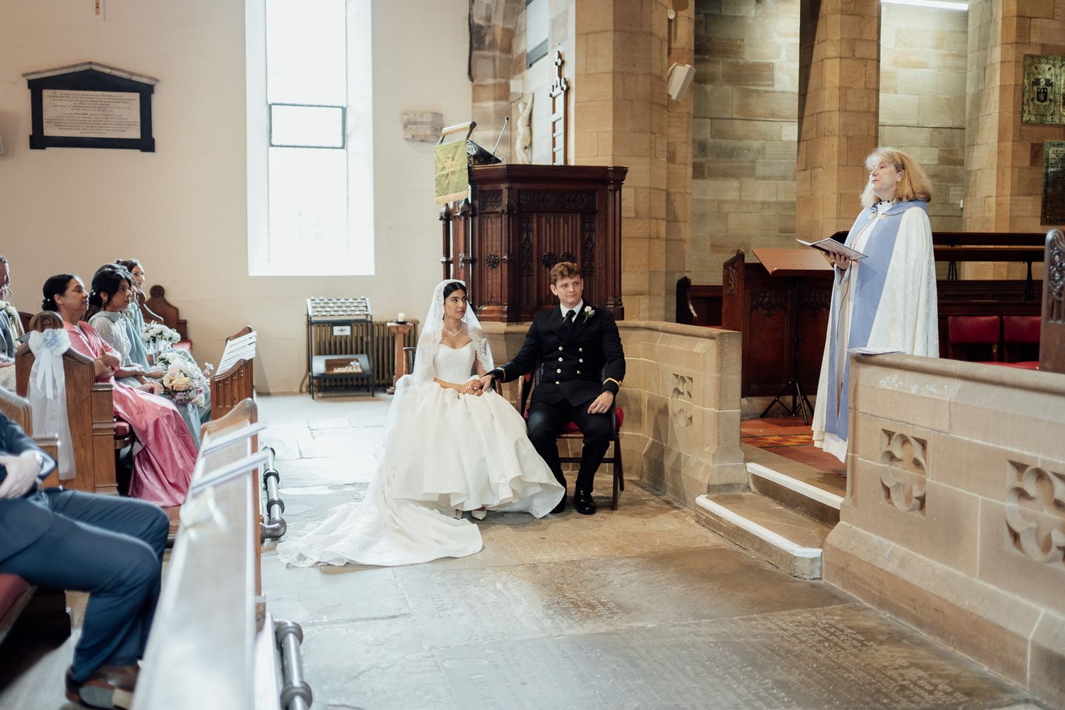 Bride and groom seated at church altar during wedding ceremony, officiant at podium, guests in pews.