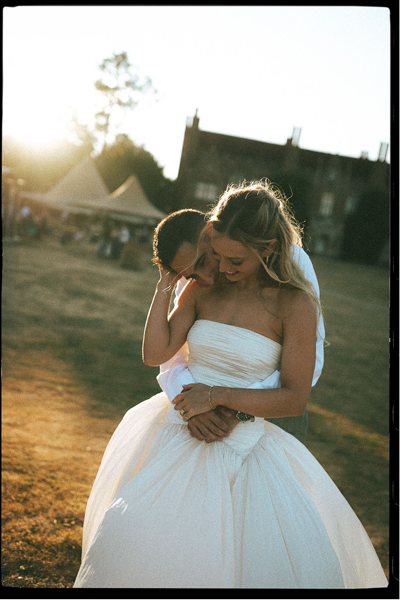 Couple embracing at golden hour wedding, bride in strapless white ball gown, warm sunlight glowing behind them.