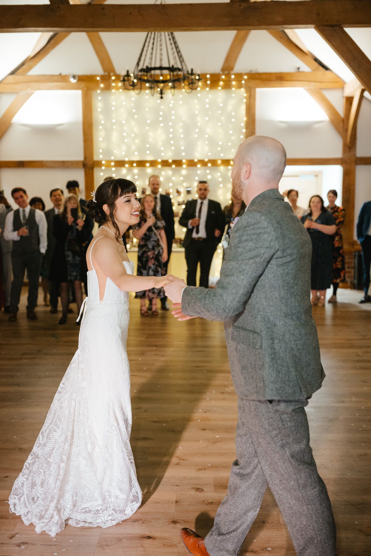 A couple shares their first dance in a rustic barn venue with string lights and wooden beams overhead.