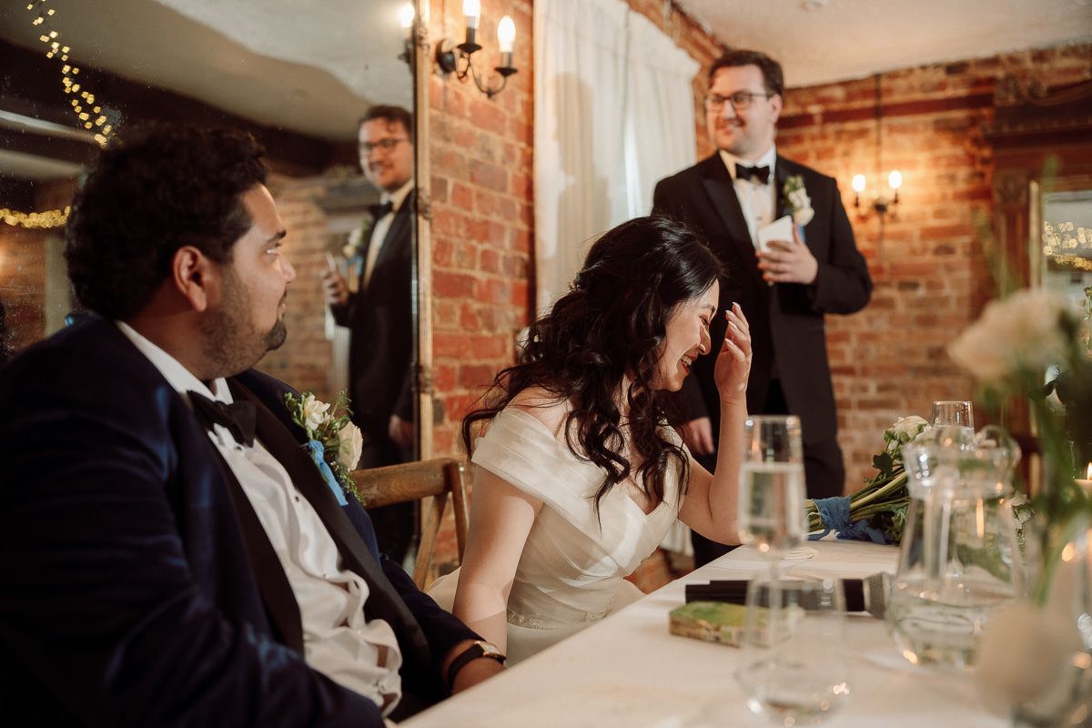 Wedding reception guests at a rustic brick-walled venue share a candid moment during dinner speeches.