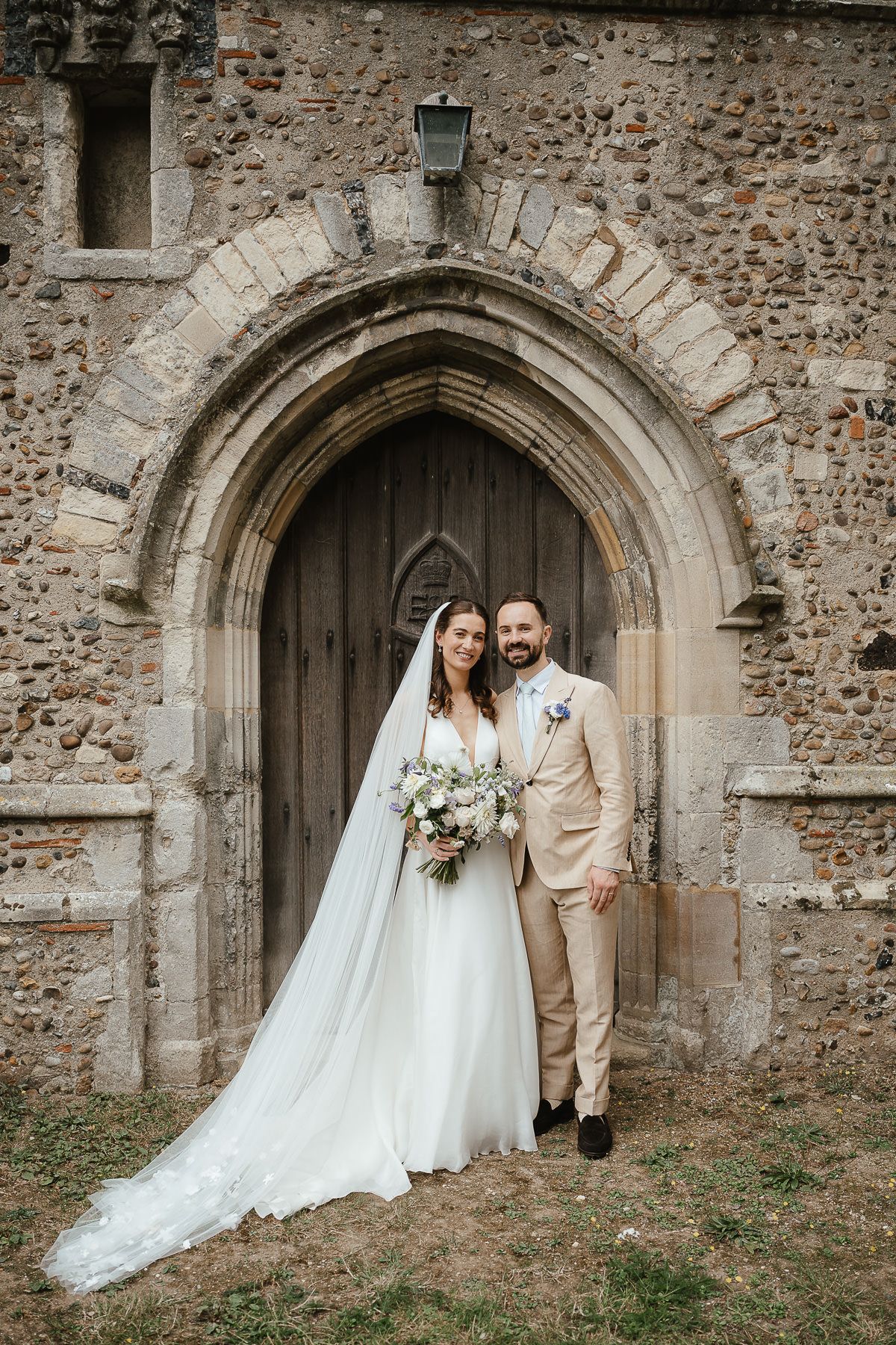 A bride and groom pose in front of a rustic stone church archway with beautiful flowing wedding dress and floral bouquet.