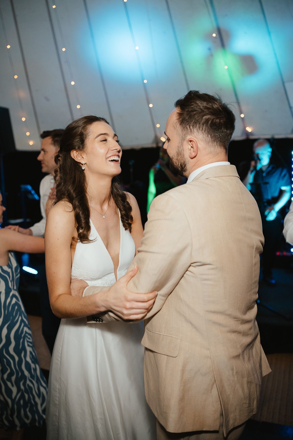 A joyful couple shares a laugh while dancing together at their wedding reception under blue mood lighting and twinkle lights.