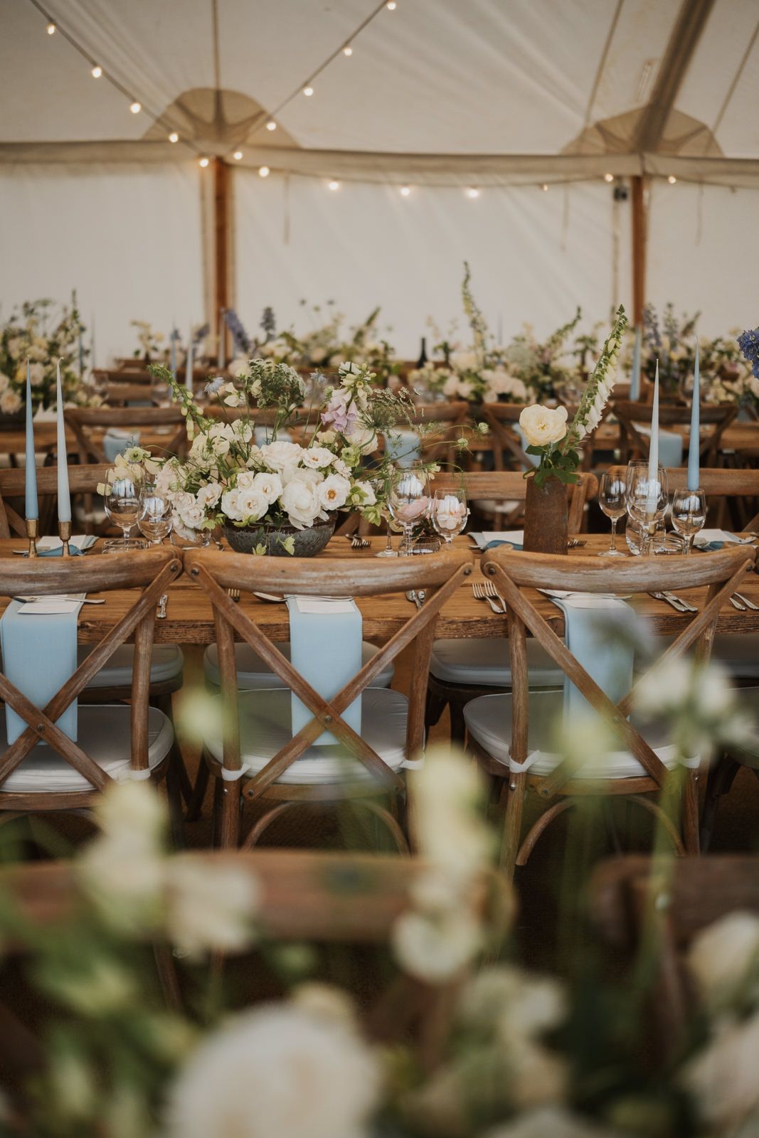 Elegant wedding reception table setting with rustic cross-back chairs and soft blue accents under white tent canopy.