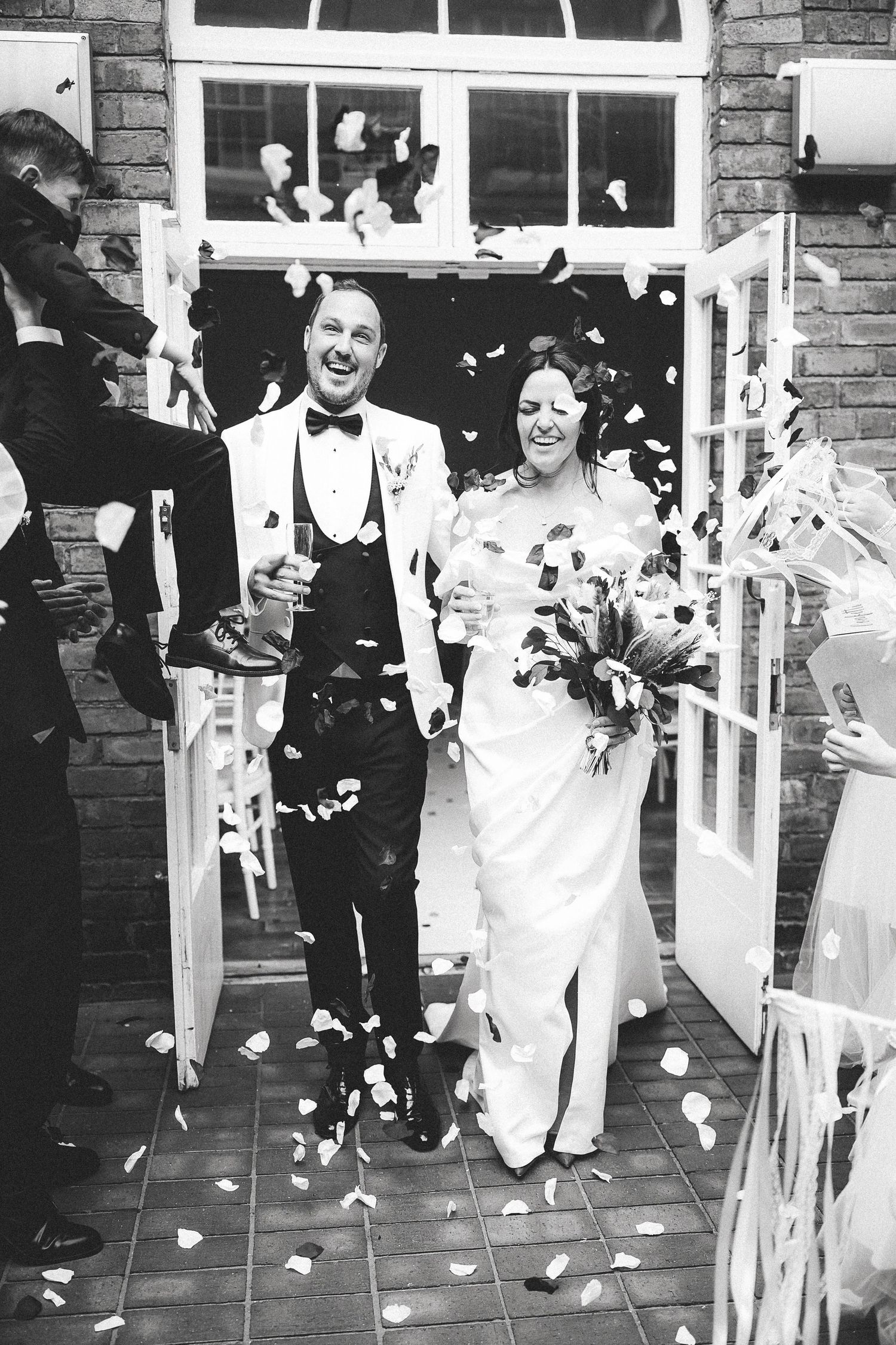 Black and white photo of a joyful newlywed couple exiting through doors while guests shower them with confetti petals.