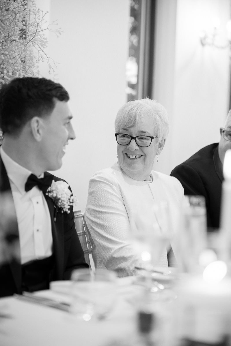 Black and white candid photo of wedding guests sharing laughter during reception dinner.