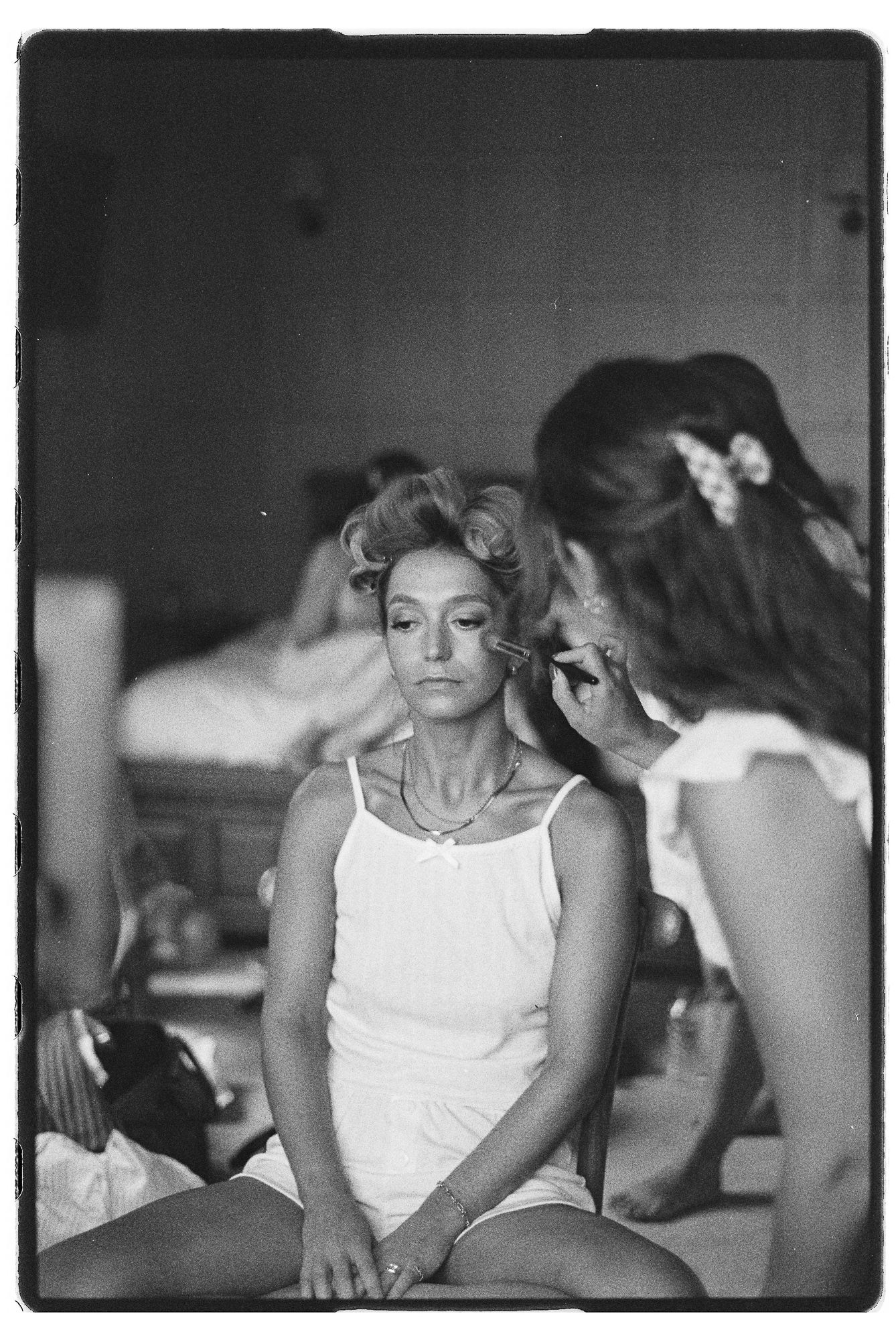 Vintage black and white photo of a woman having makeup applied while sitting, with another person visible in background.