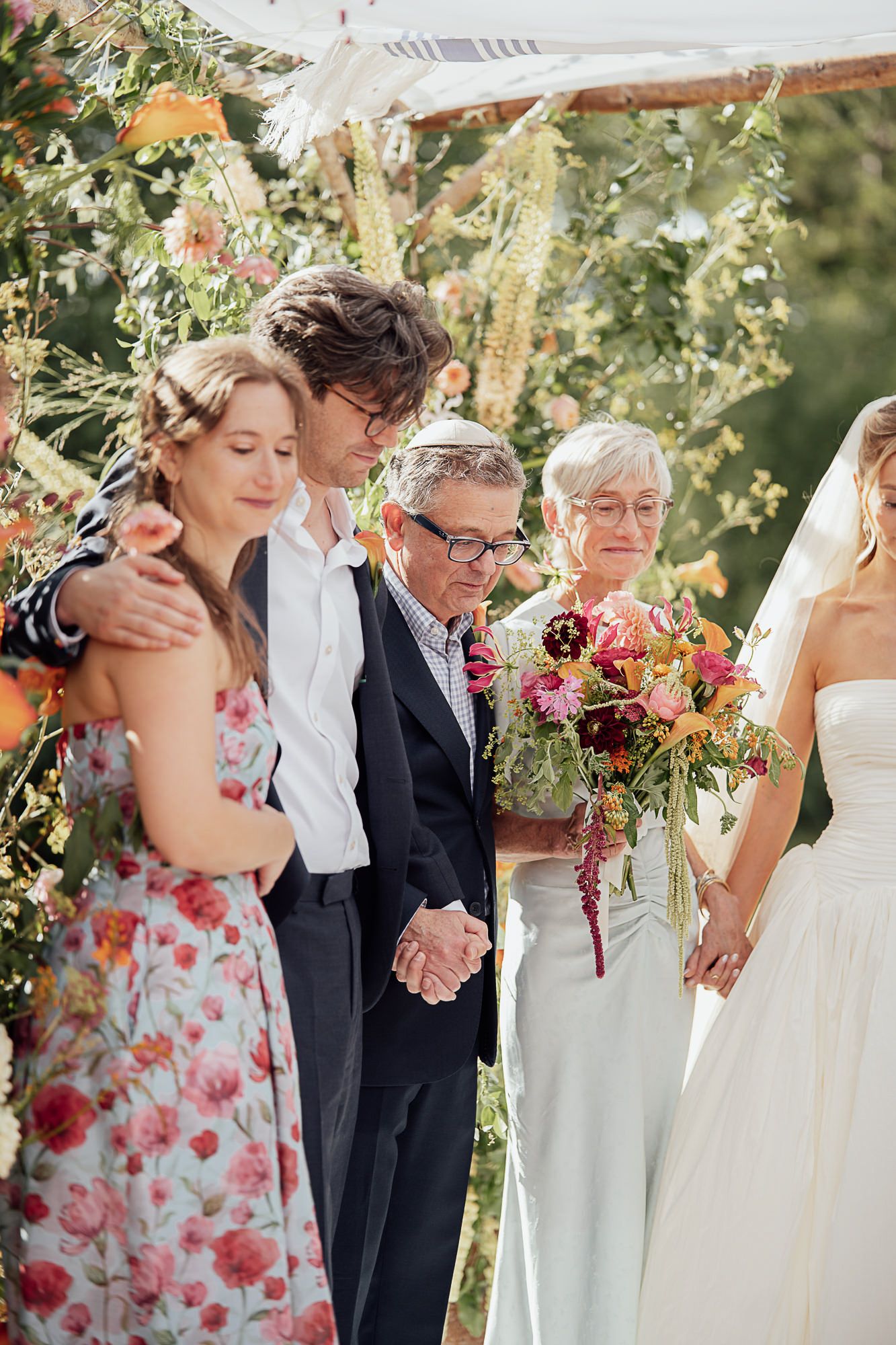 Family gathering at an outdoor wedding ceremony with colorful floral arch, bride holding bouquet.
