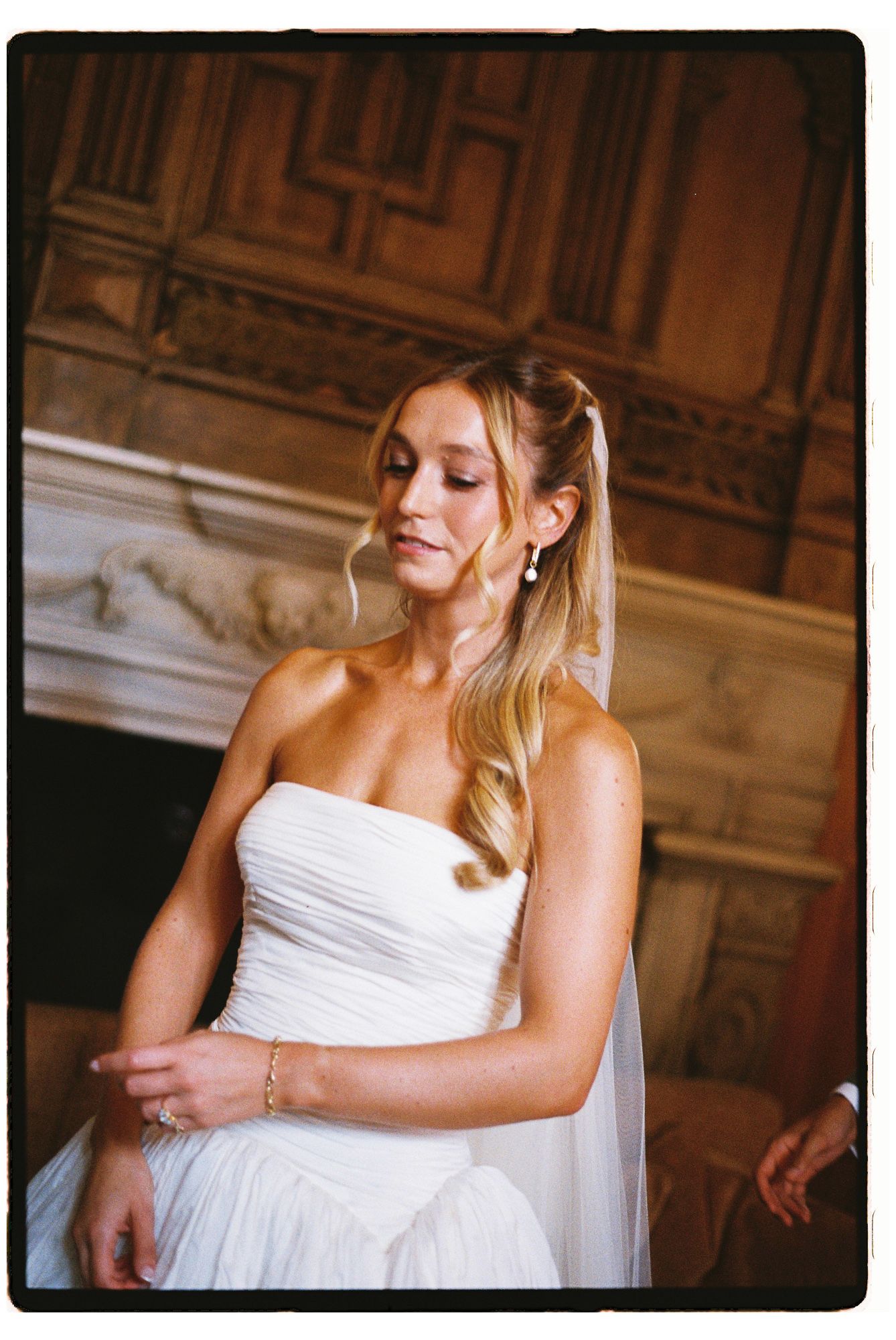 Bride in strapless white ruched gown with veil, looking down elegantly before ornate wood fireplace.