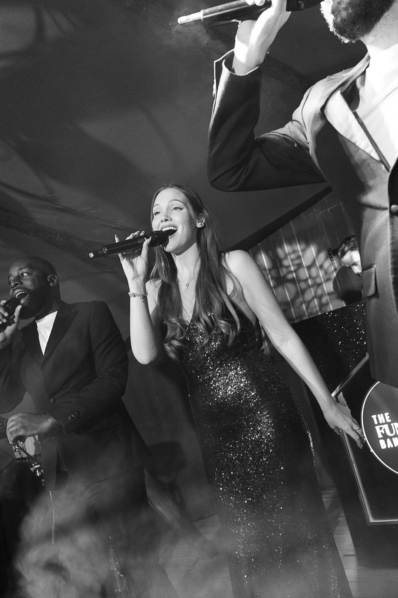 Black and white photo of a woman in a sequin dress singing into a microphone on stage with band members behind her.