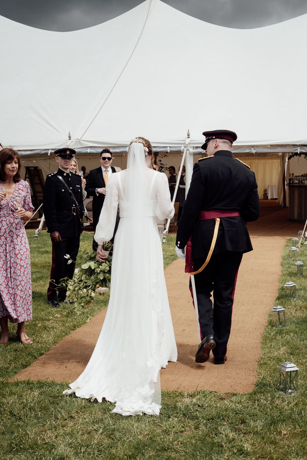 A wedding party walks through a large white tent during an outdoor reception on grassy grounds.