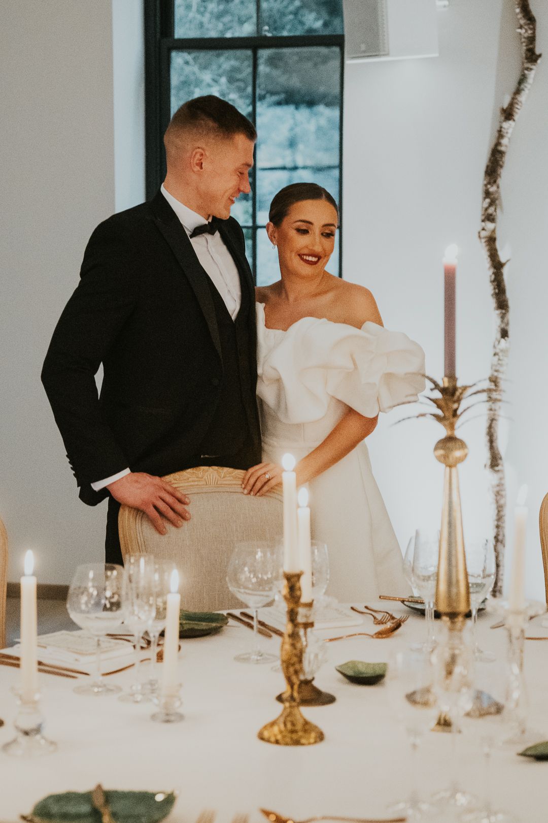 Elegant newlywed couple stands at candlelit reception table decorated with gold tapered candles and place settings.