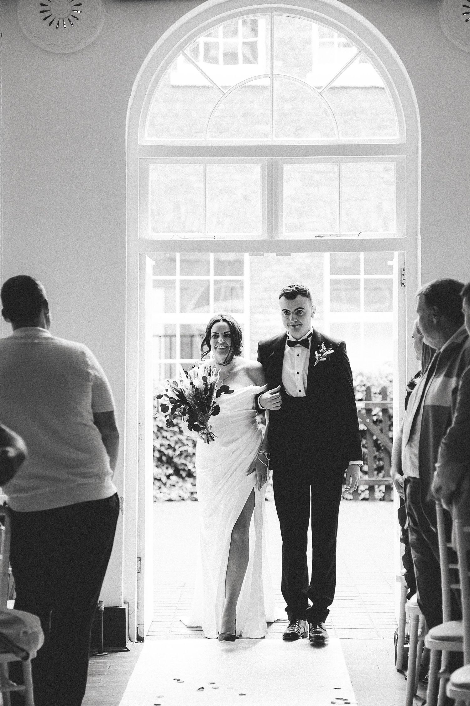 A black and white wedding ceremony photo shows silhouettes of guests standing in front of bright arched windows.