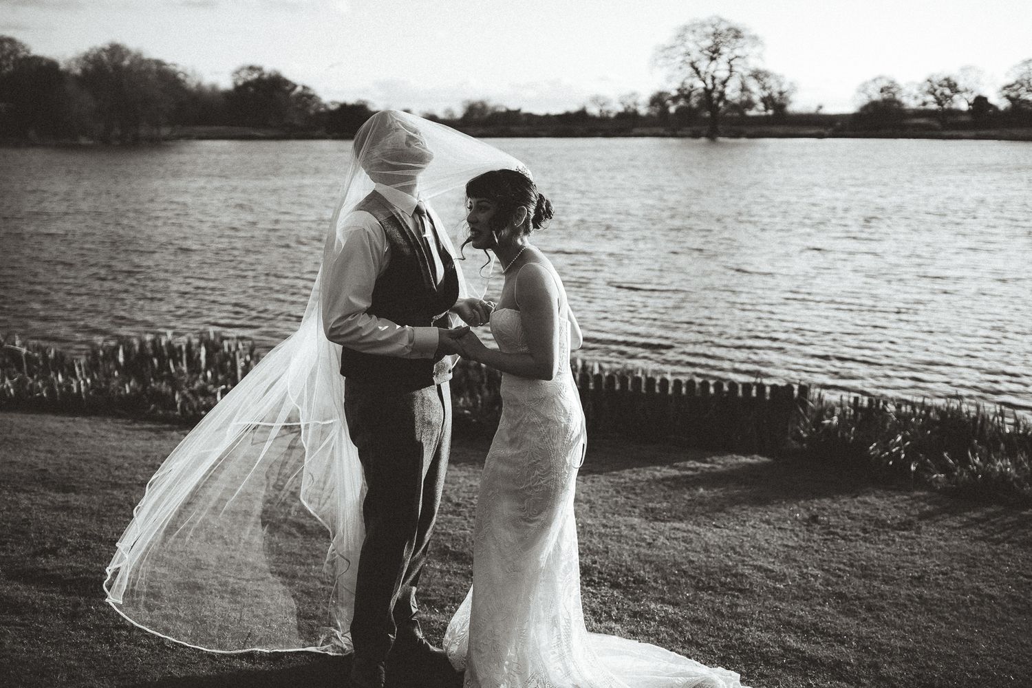 A romantic black and white wedding photo of a couple embracing by a serene lake with a flowing veil catching the light.