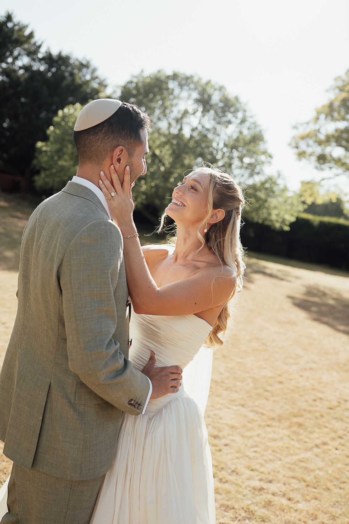 Smiling bride in white dress gazes at groom in grey suit and kippah outdoors in golden sunlight.