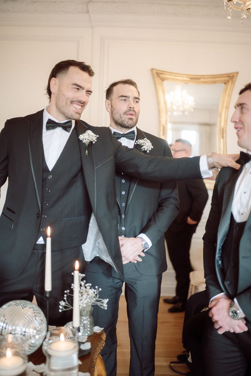 Groomsmen in black tuxedos share a candid moment while laughing together at an elegant indoor wedding venue.