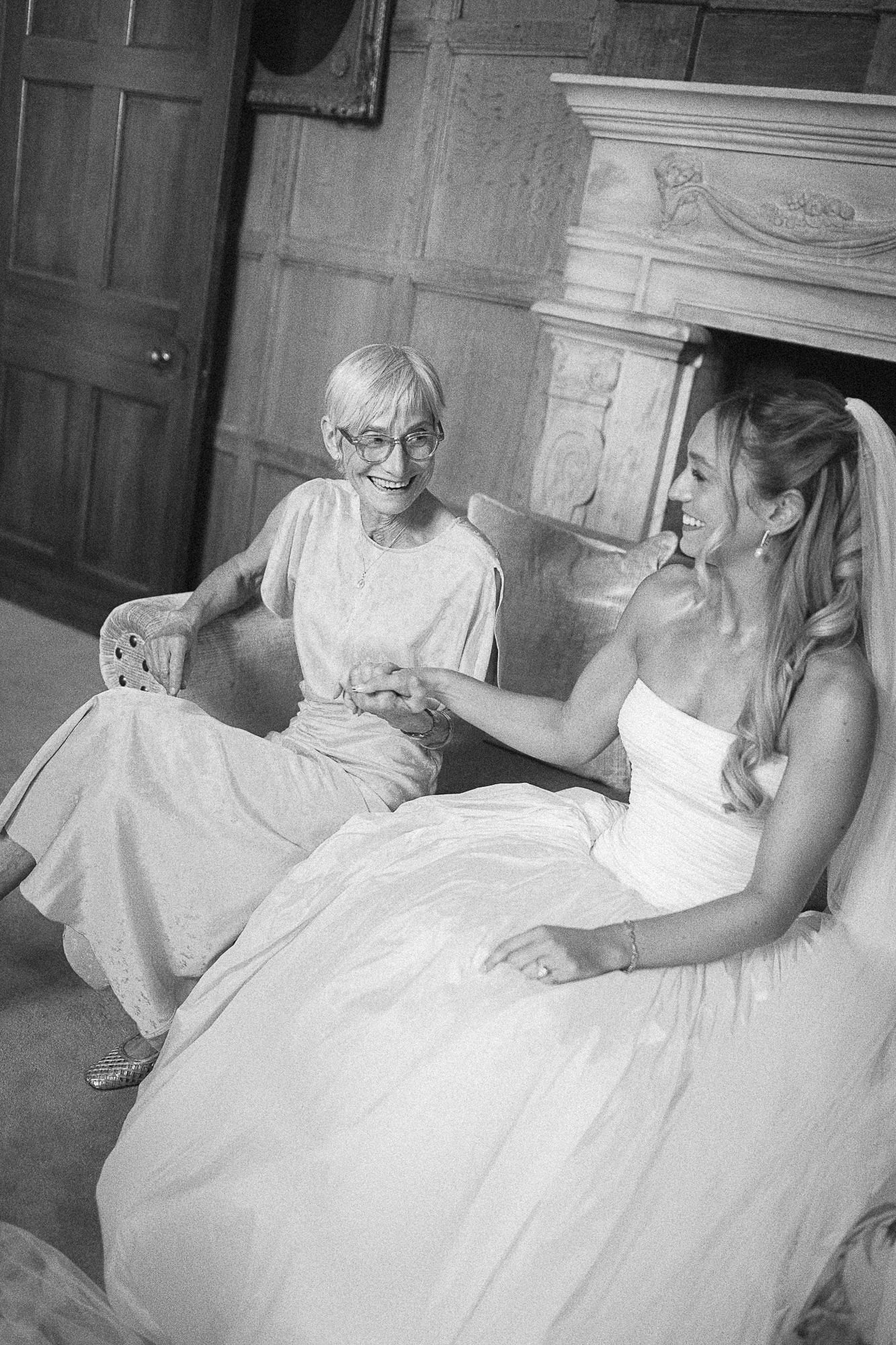 Bride in white gown holds hands with elderly woman in black and white wedding photo.