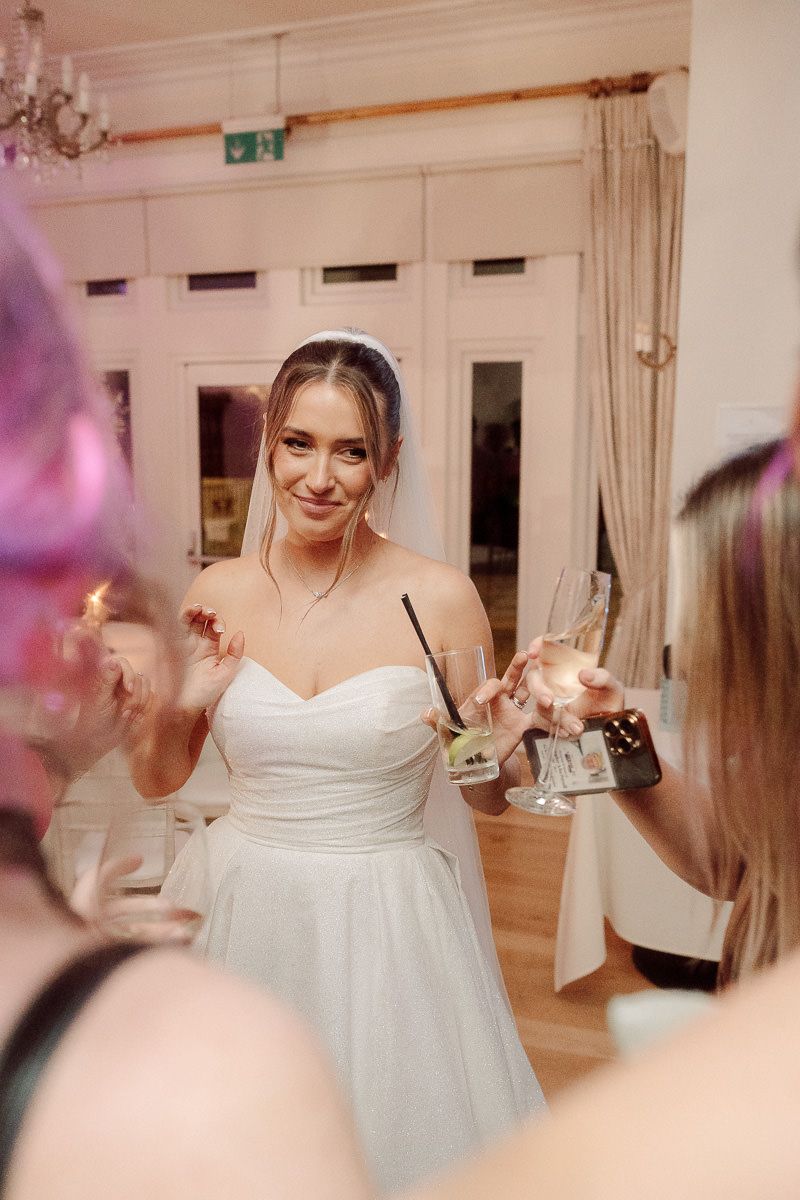 A bride in a white strapless wedding dress raises her glass during a celebration at an indoor venue.