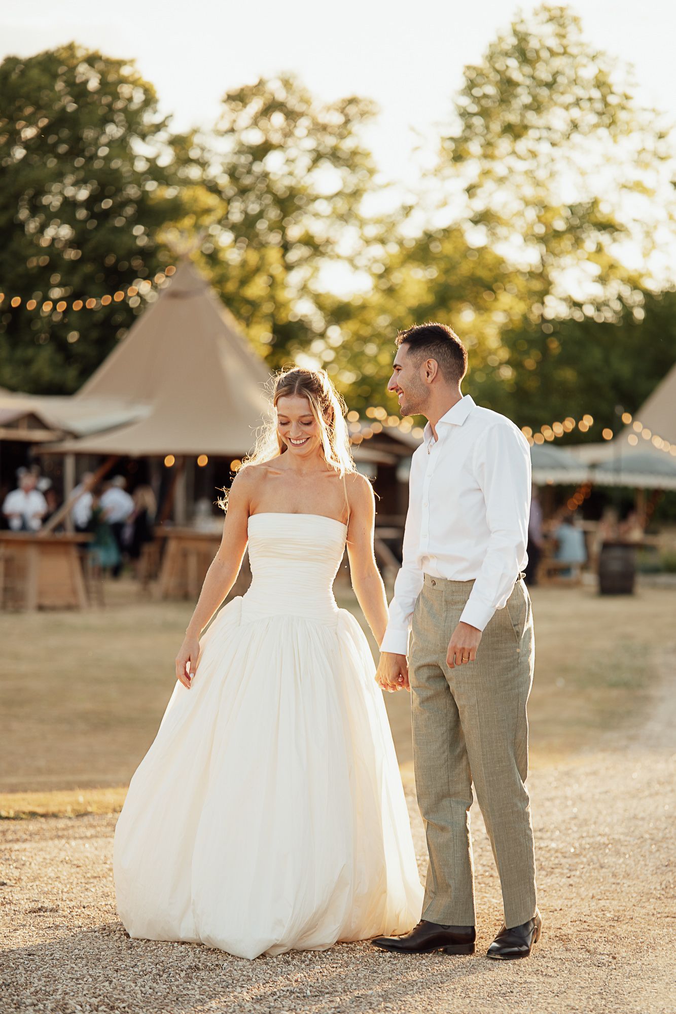 Bride and groom holding hands at golden hour outdoor wedding venue with tipi and string lights.