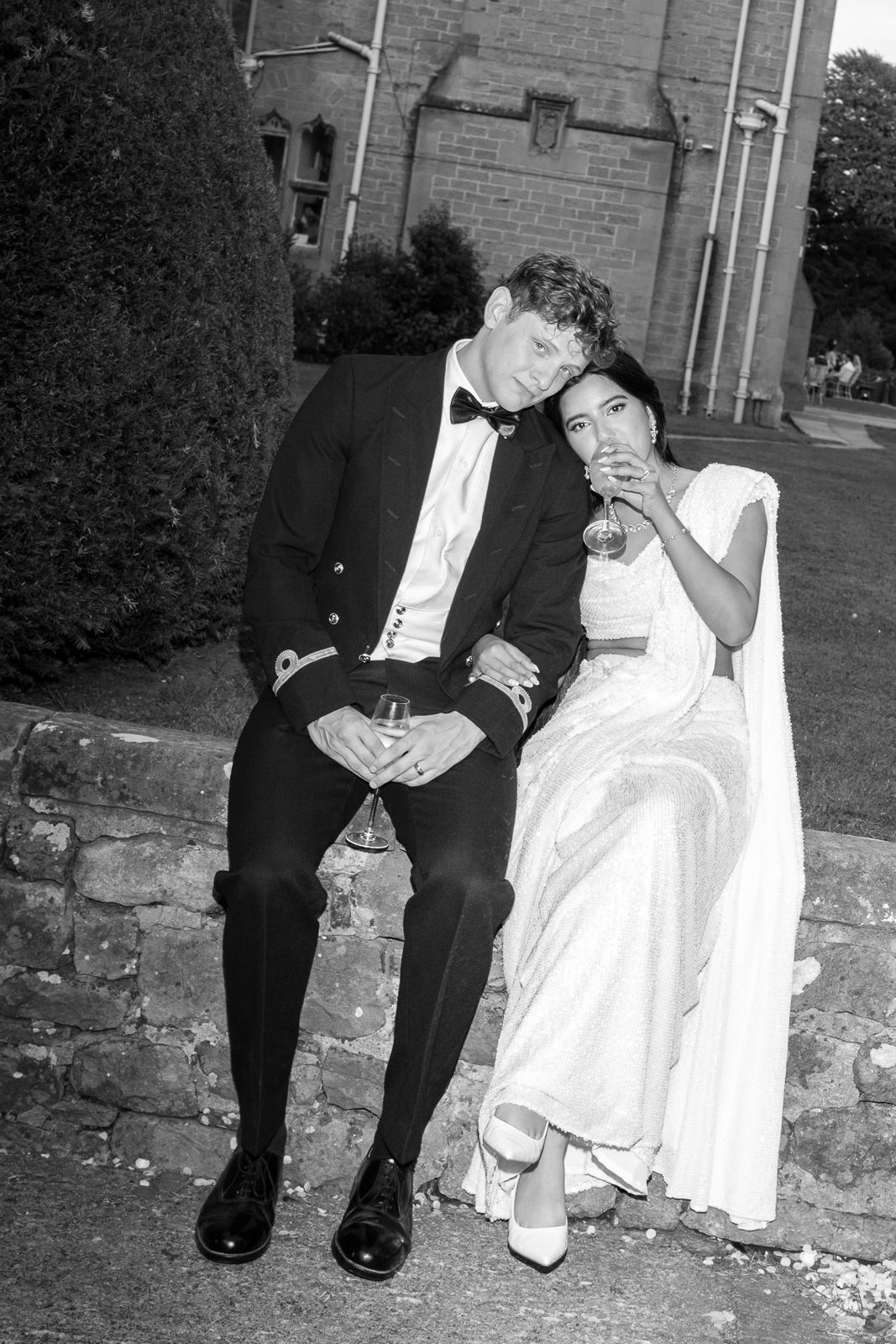 Black and white photo of bride and groom sitting on stone wall sharing drinks outside a church at dusk.