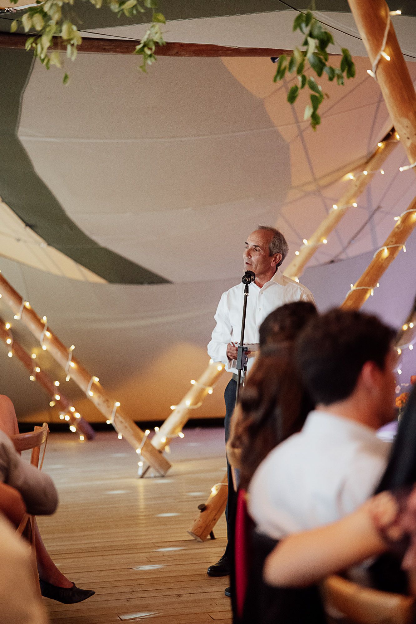 Man in white shirt giving a speech at an elegant wedding reception in a decorated tipi tent with fairy lights.