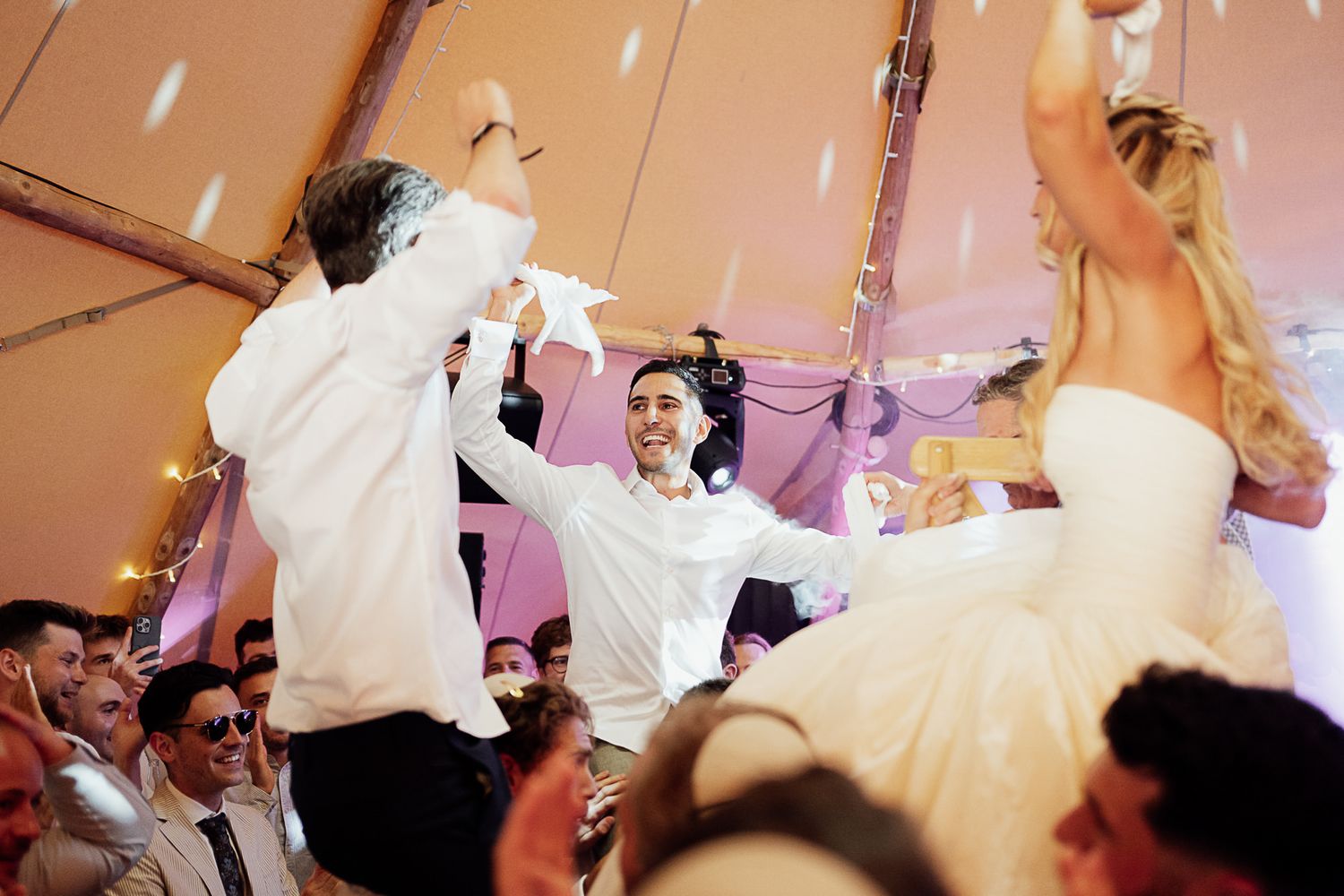 Joyful wedding reception dance under a tent with bride in white gown and guests celebrating with raised hands.