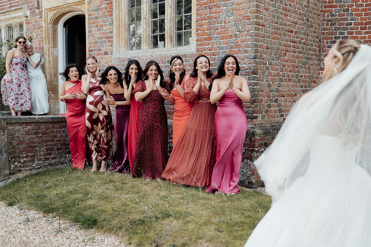 Bridesmaids in red and pink dresses react with joy as bride in white gown approaches brick manor.