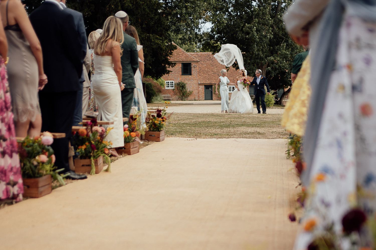 Outdoor wedding ceremony aisle lined with colorful floral arrangements, guests standing as couple exchanges vows near rustic barn.