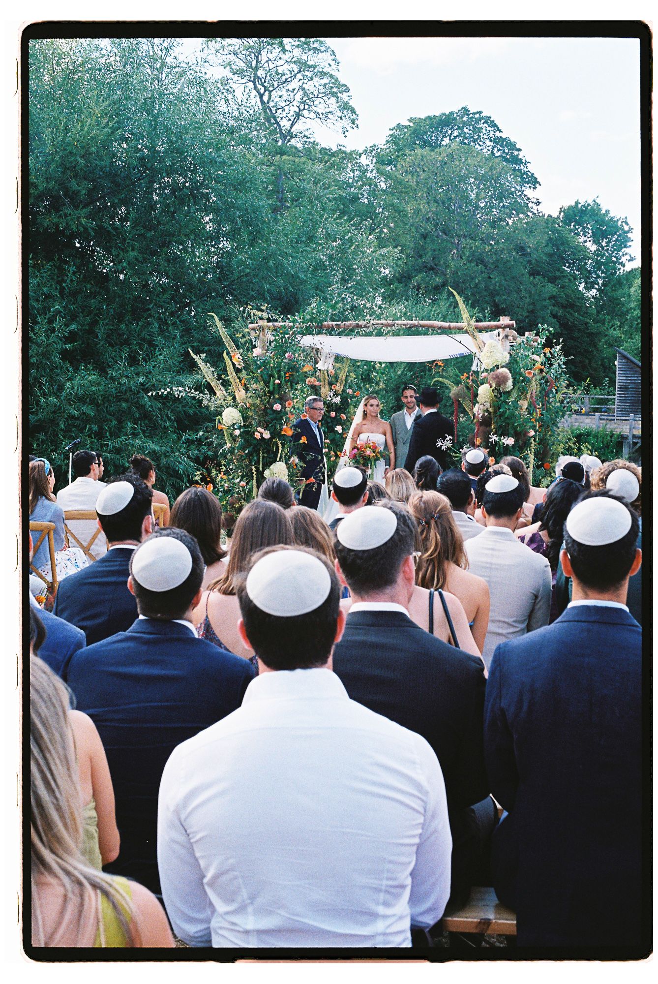 Outdoor Jewish wedding ceremony with guests wearing kippot, couple under floral chuppah, surrounded by lush greenery.