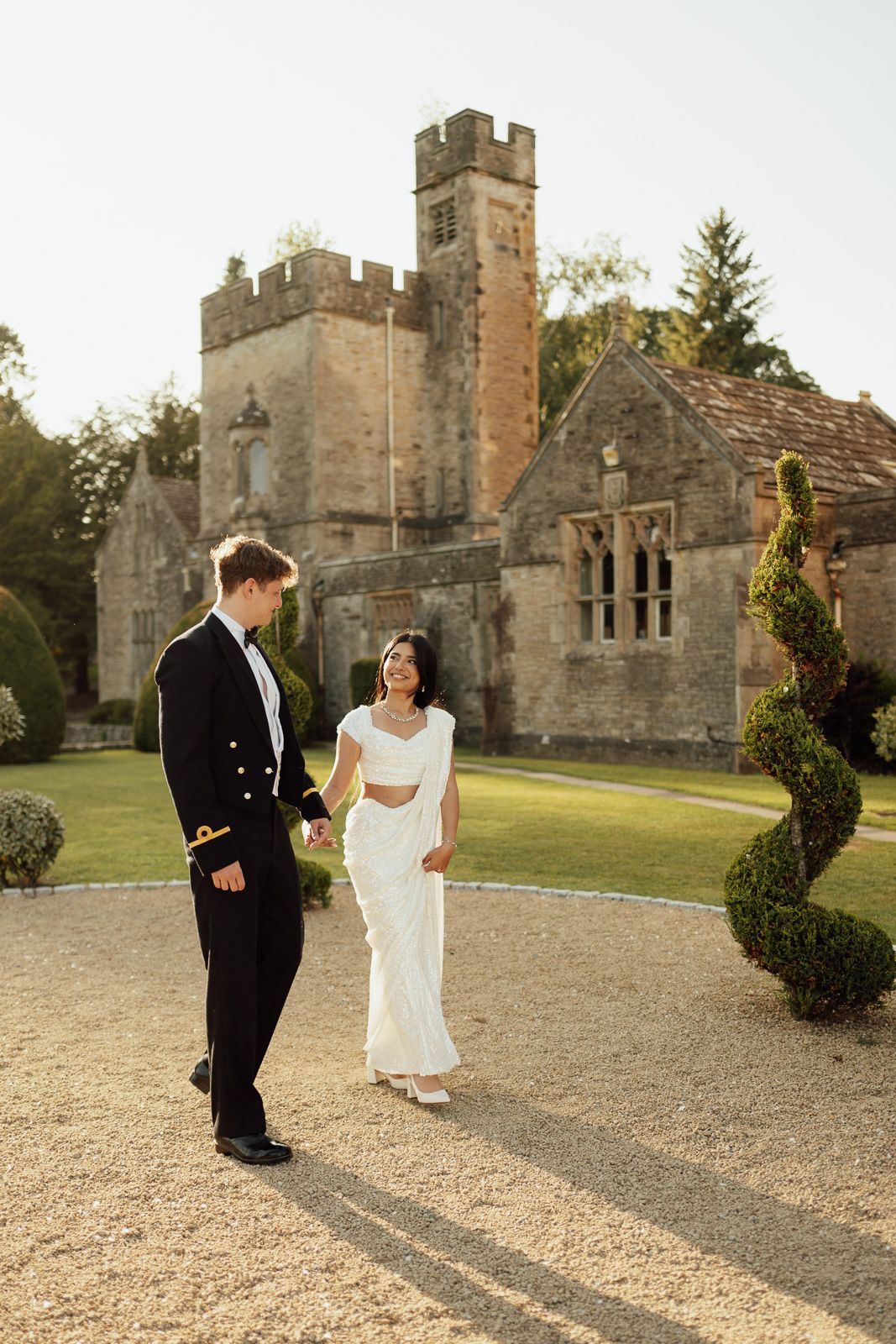 Bride and groom walking hand in hand on gravel path before a historic stone manor in golden hour light.