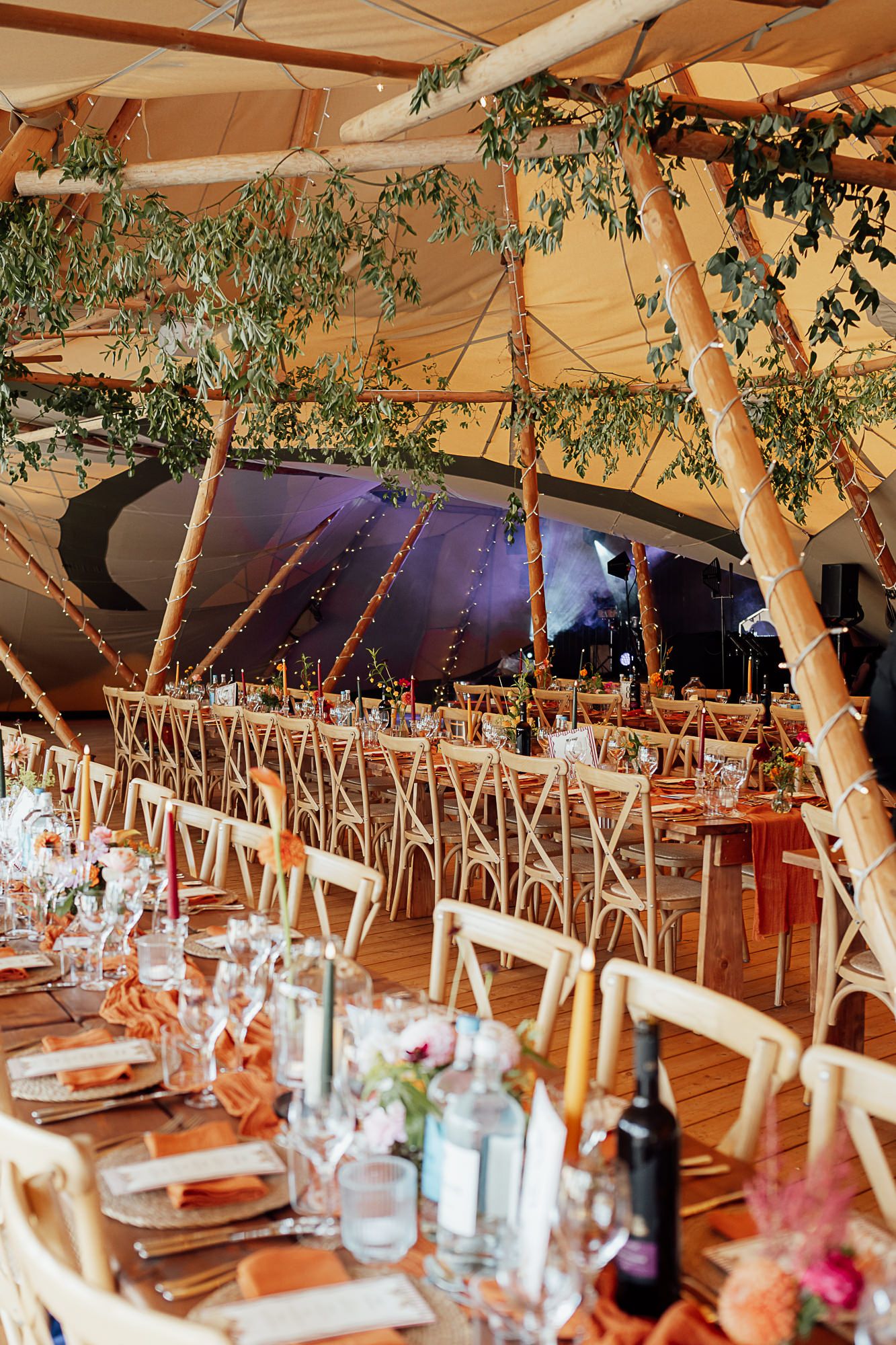 Elegant tipi wedding reception with long dining tables, cross-back chairs, greenery decor, and warm string lights.