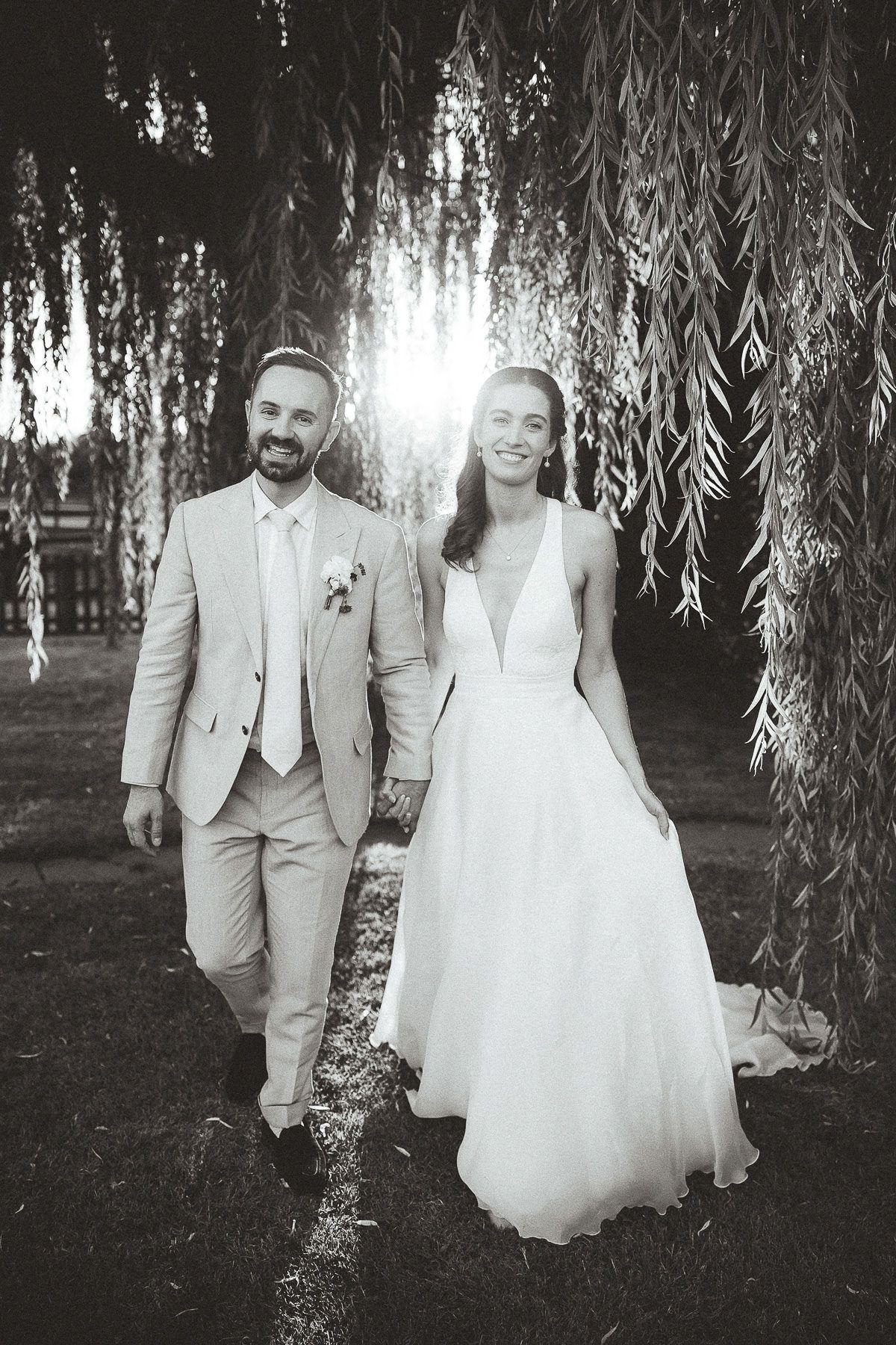 Bride and groom walk together under romantic drooping willow trees in a garden setting.