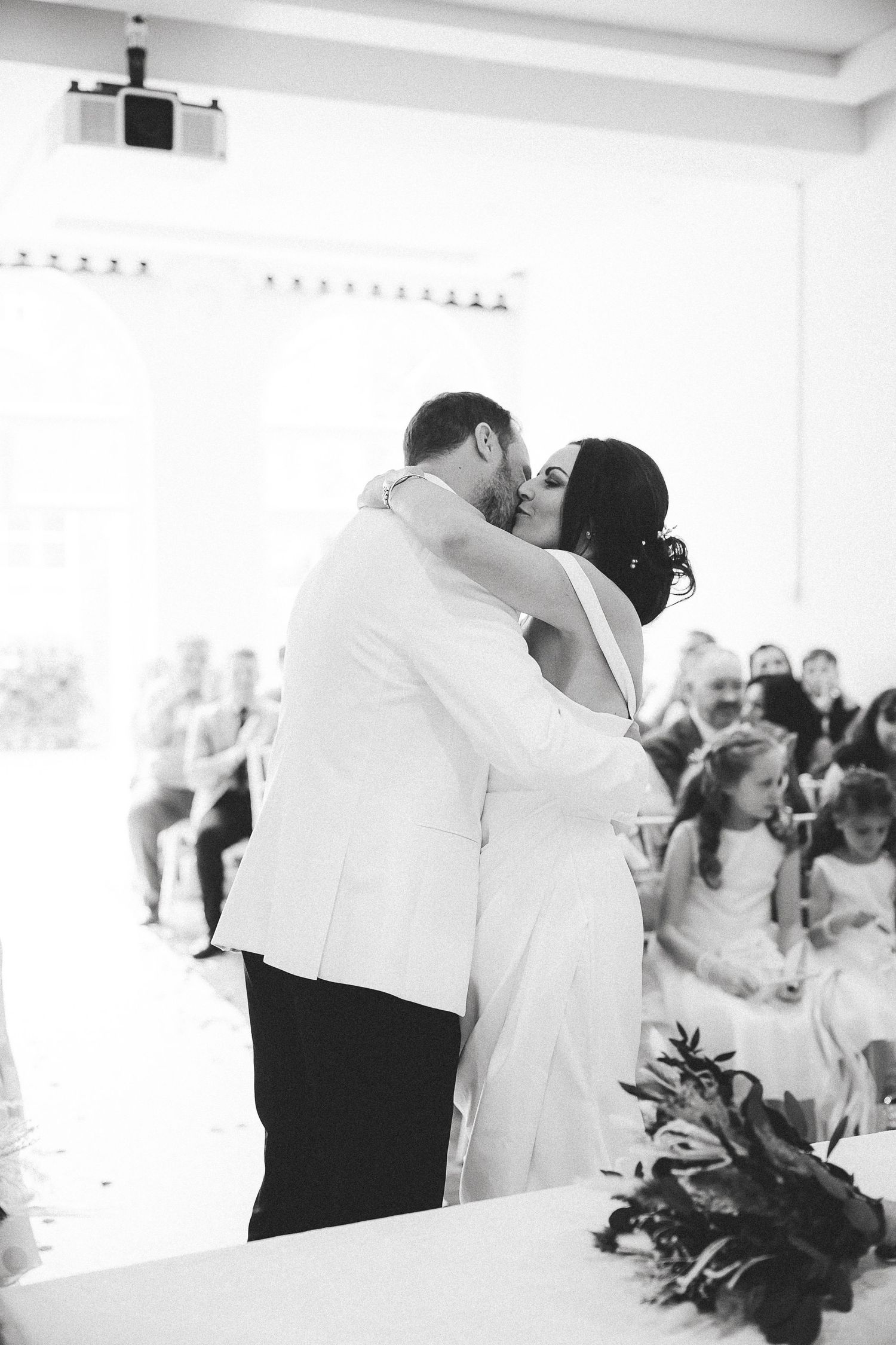 A romantic black and white photo captures a couple sharing their first kiss during their wedding ceremony.