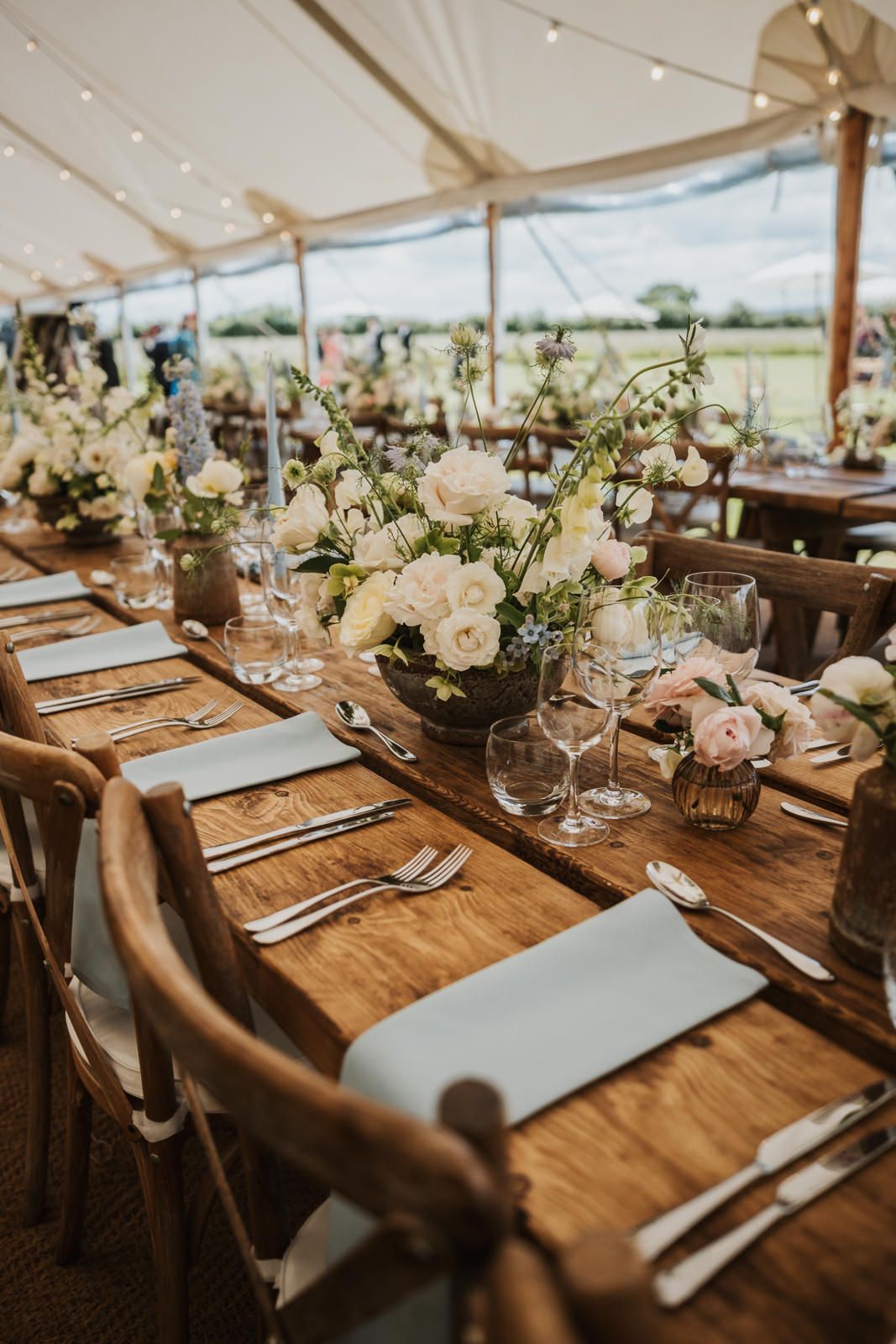 Rustic outdoor wedding reception table setting with white floral centerpieces and wooden chairs under an elegant tent canopy.