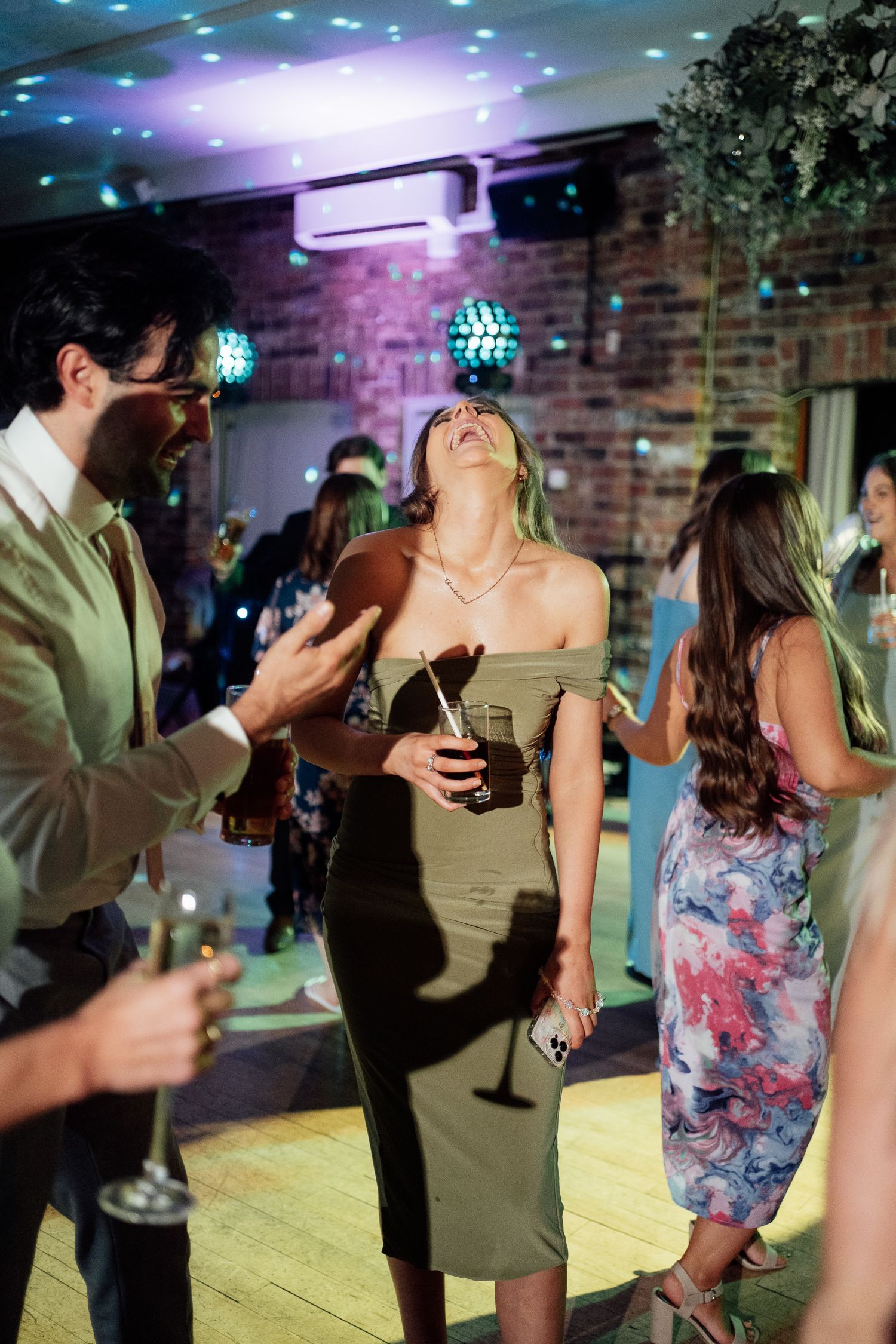 Party guests laugh and dance together at a social event with disco lights illuminating the brick wall backdrop.