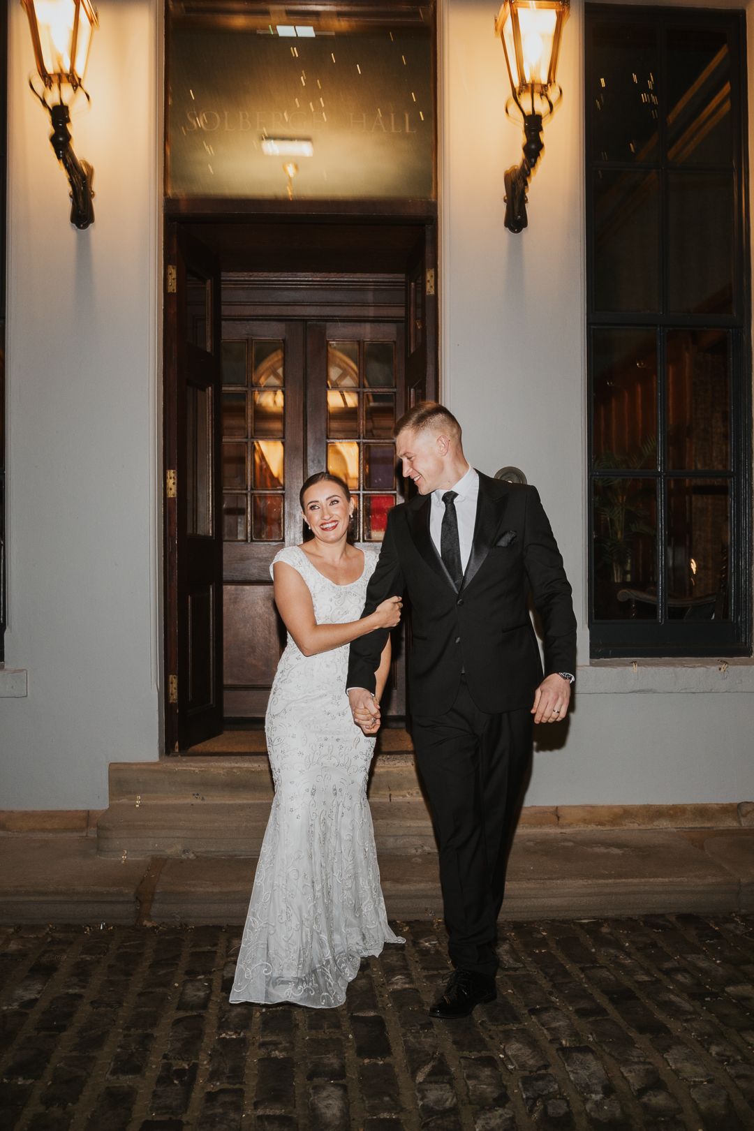 A wedding couple walks together along a cobblestone path outside an elegant venue with wall sconces at night.