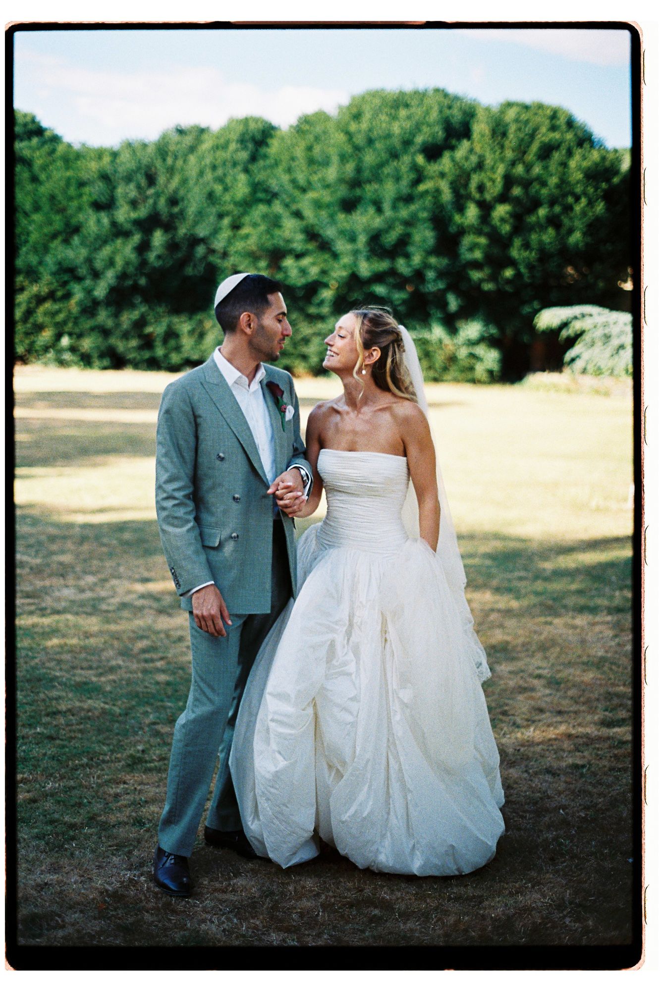 Bride in strapless white ballgown and groom in gray suit share a romantic moment outdoors on a sunny wedding day.