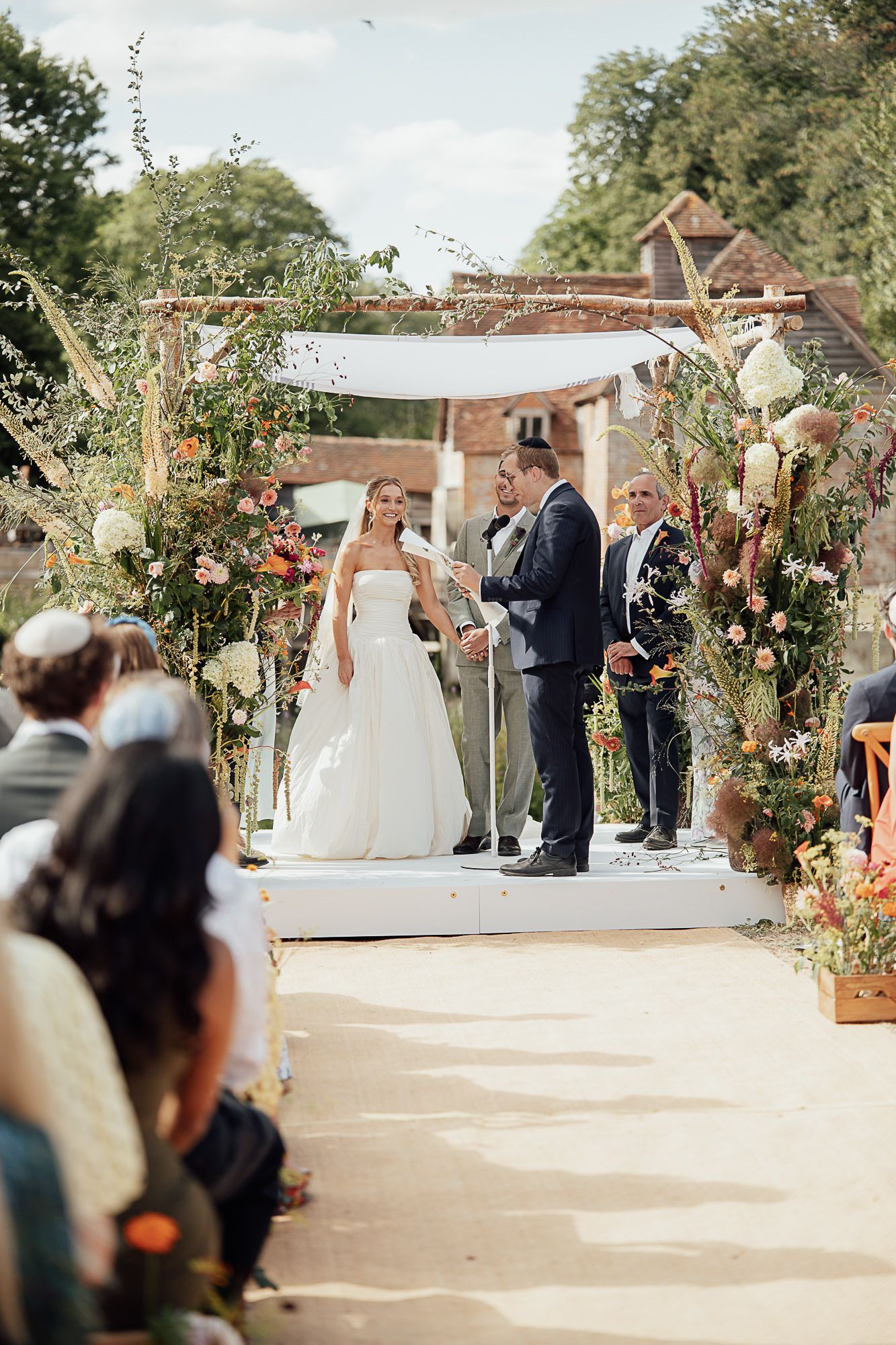Bride and groom exchange vows under a floral arch at an outdoor wedding ceremony with guests seated on a sunny day.